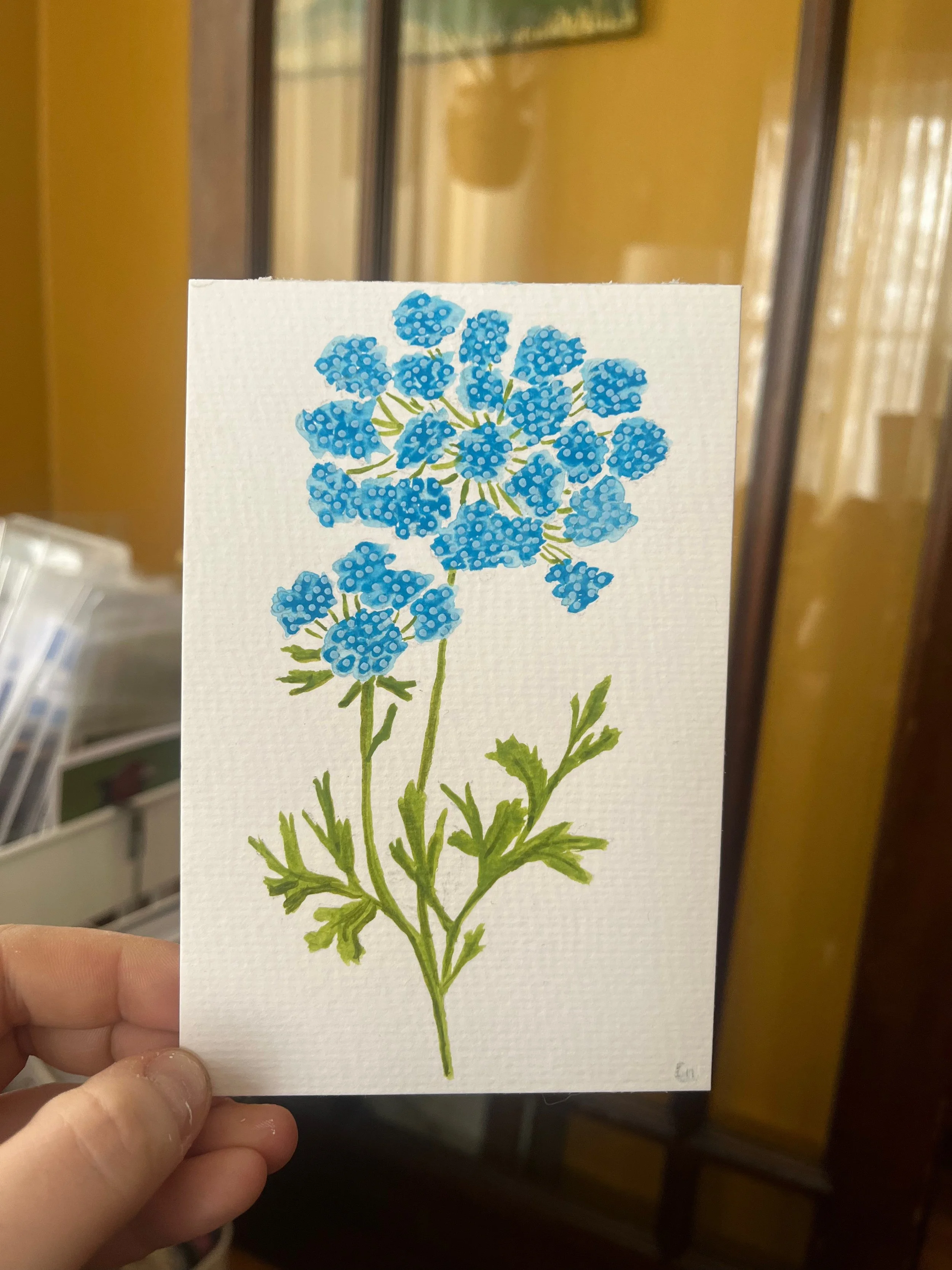 Hand holding a watercolor painting of a bouquet of blue flowers with green leaves on white textured paper.
