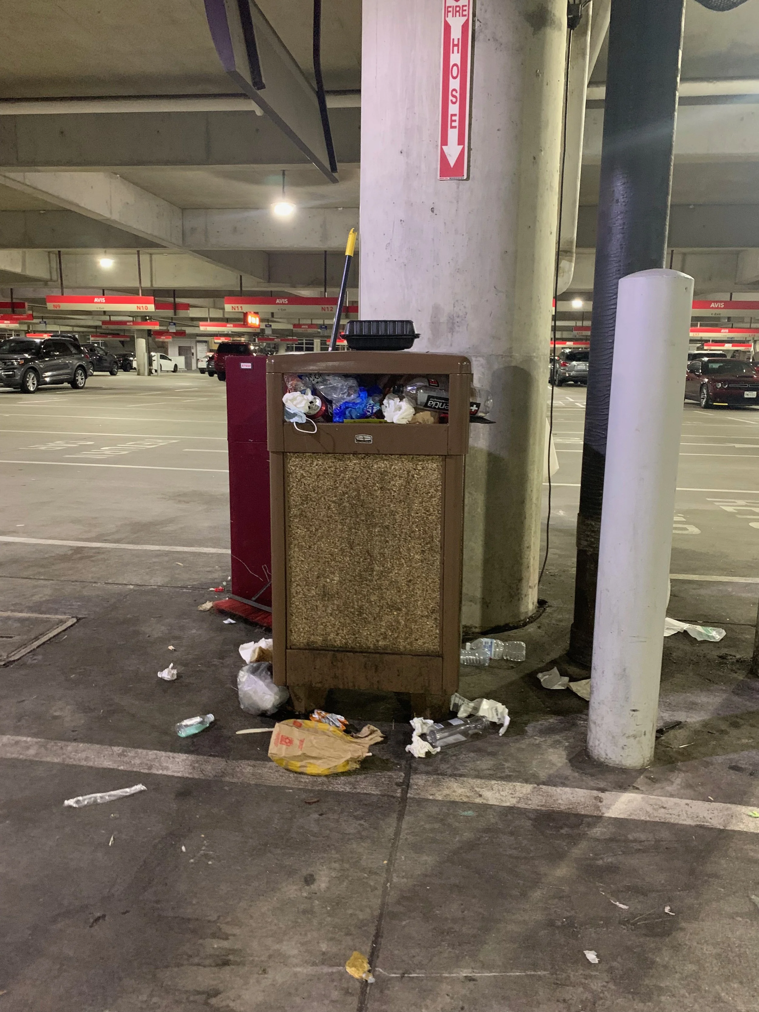 Overflowing trash bin in a parking garage with litter on the ground around it, including crumpled paper, plastic bags, and empty bottles.