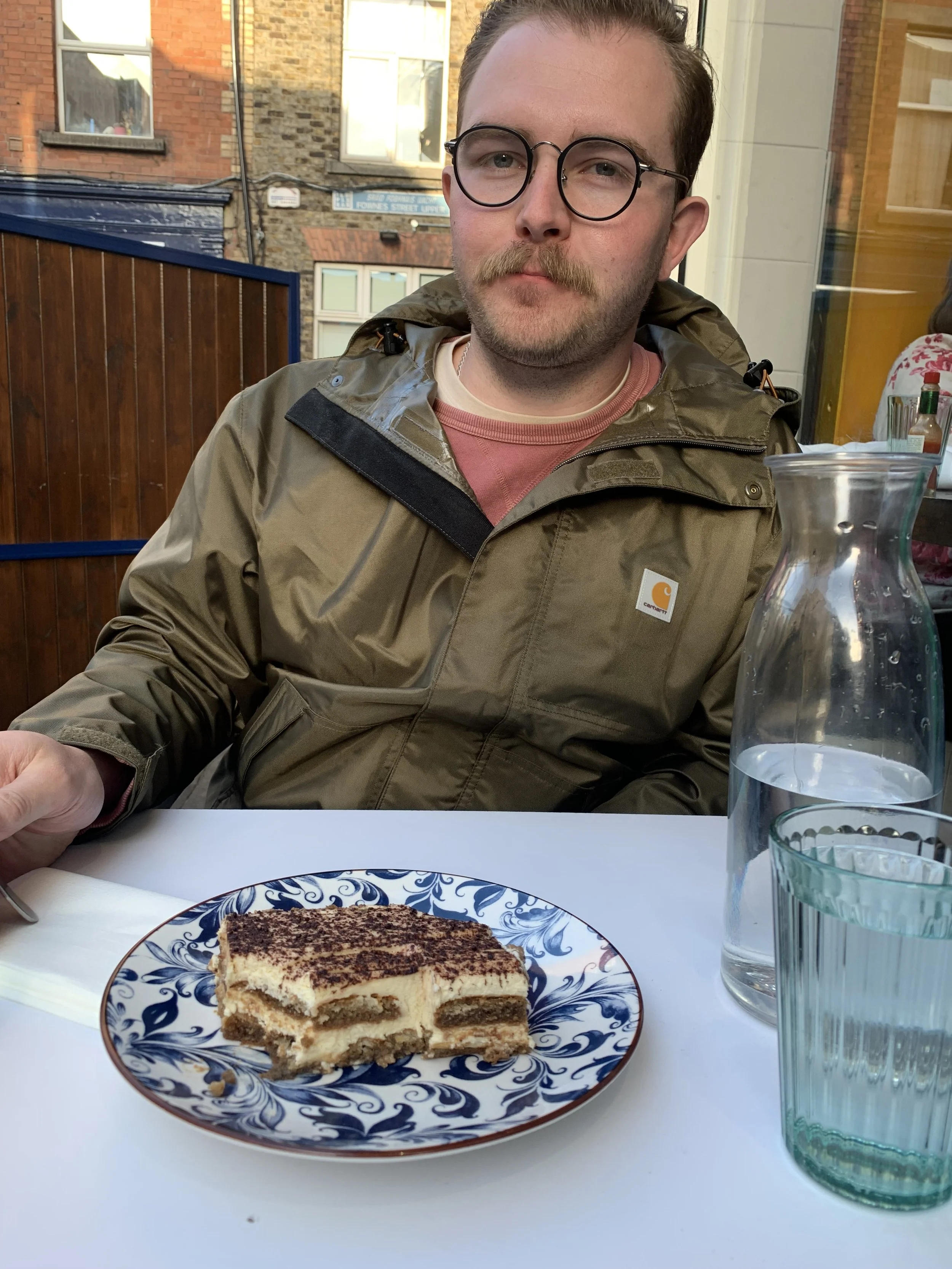 A man wearing glasses and a brown Carhartt jacket sitting at a table with a slice of tiramisu on a decorative blue and white plate, a glass pitcher of water, and a glass of water, with an outdoor view of buildings in the background.