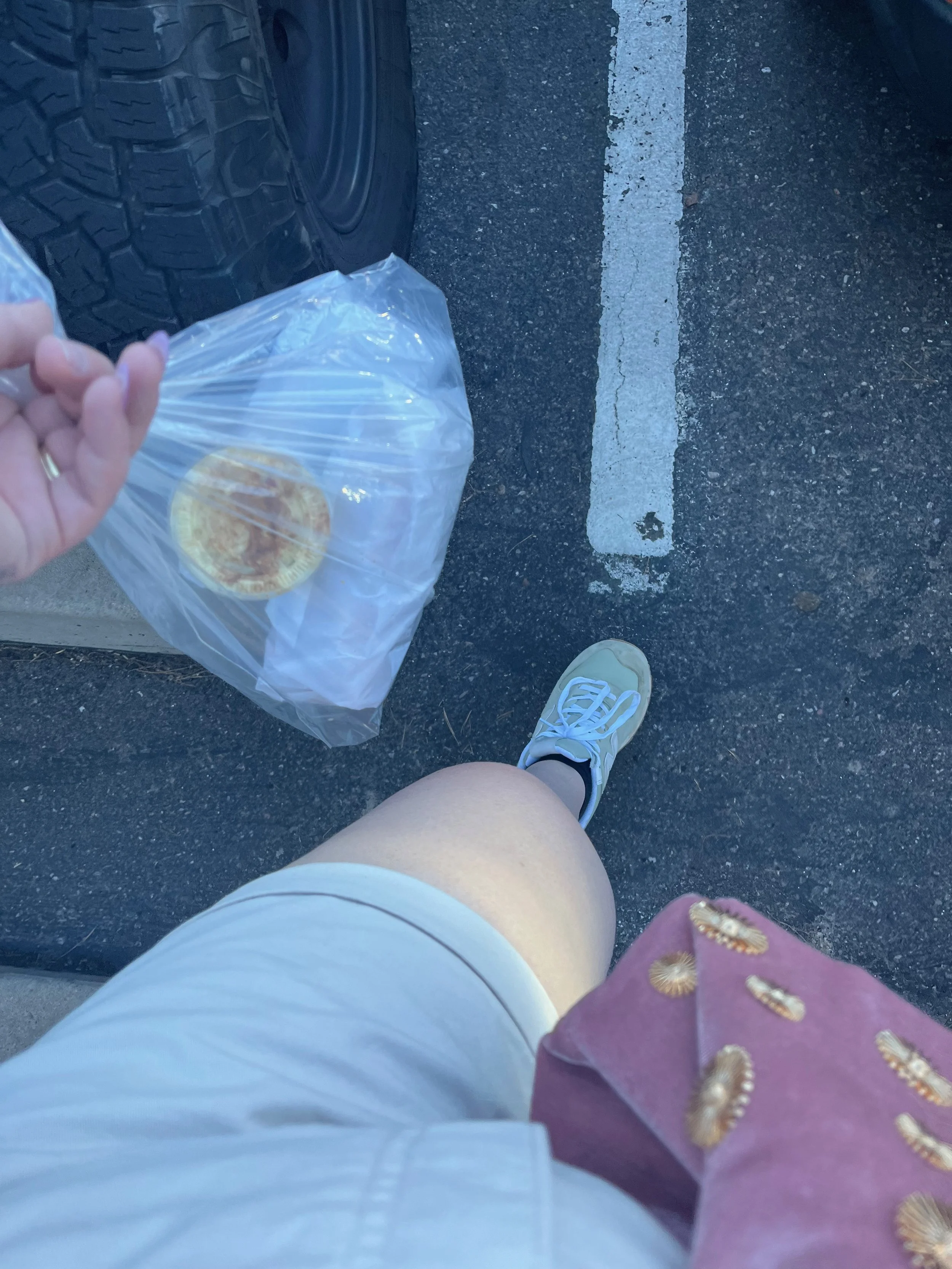 A person's legs, wearing white shorts and sneakers, standing in a parking lot with a plastic bag hanging from their hand, which contains a burger. The parking lot has a black asphalt surface with a white line.