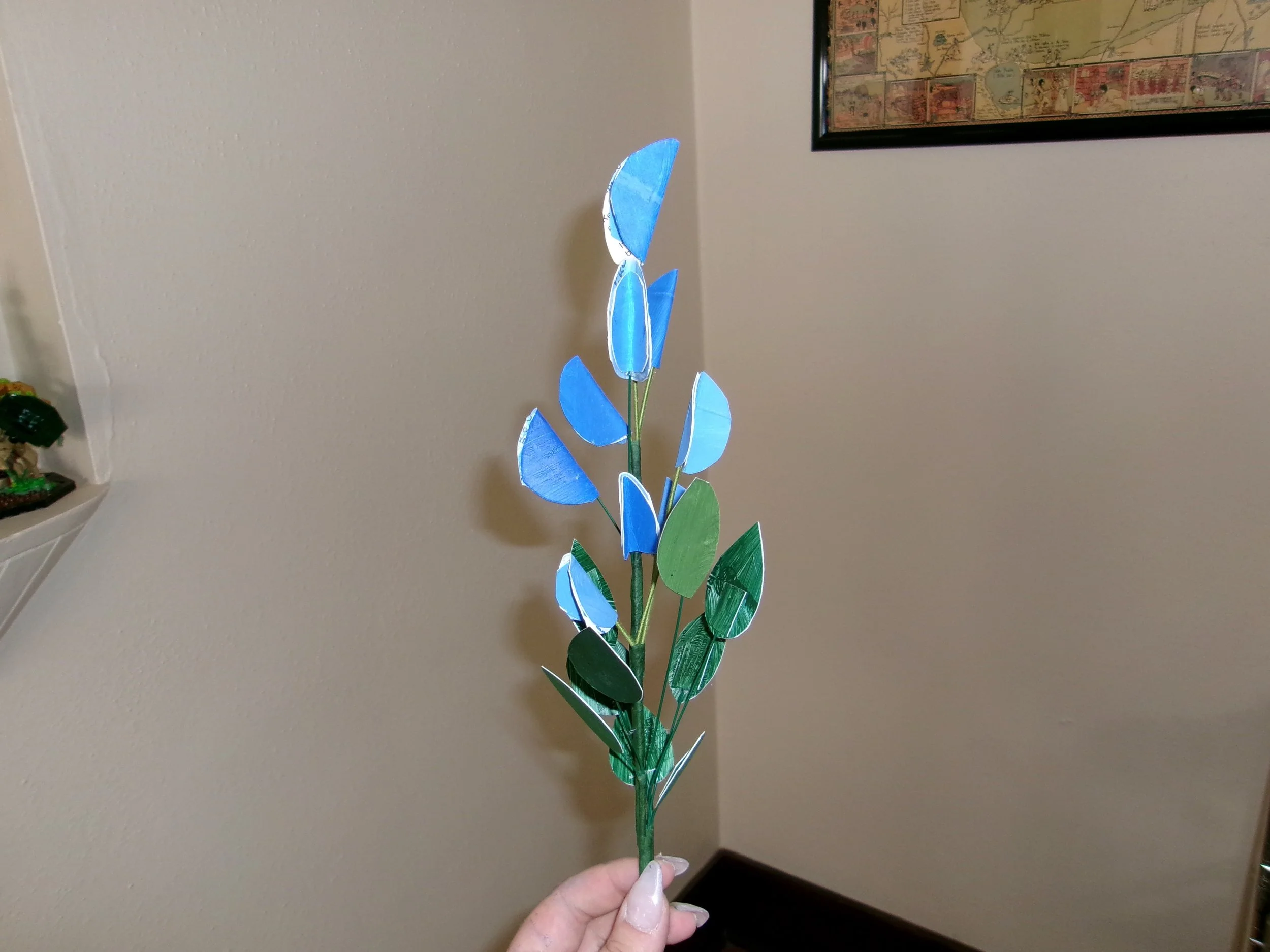 A hand holding a bouquet of paper-made blue and green leaves and flowers against a beige indoor wall with framed artwork visible.