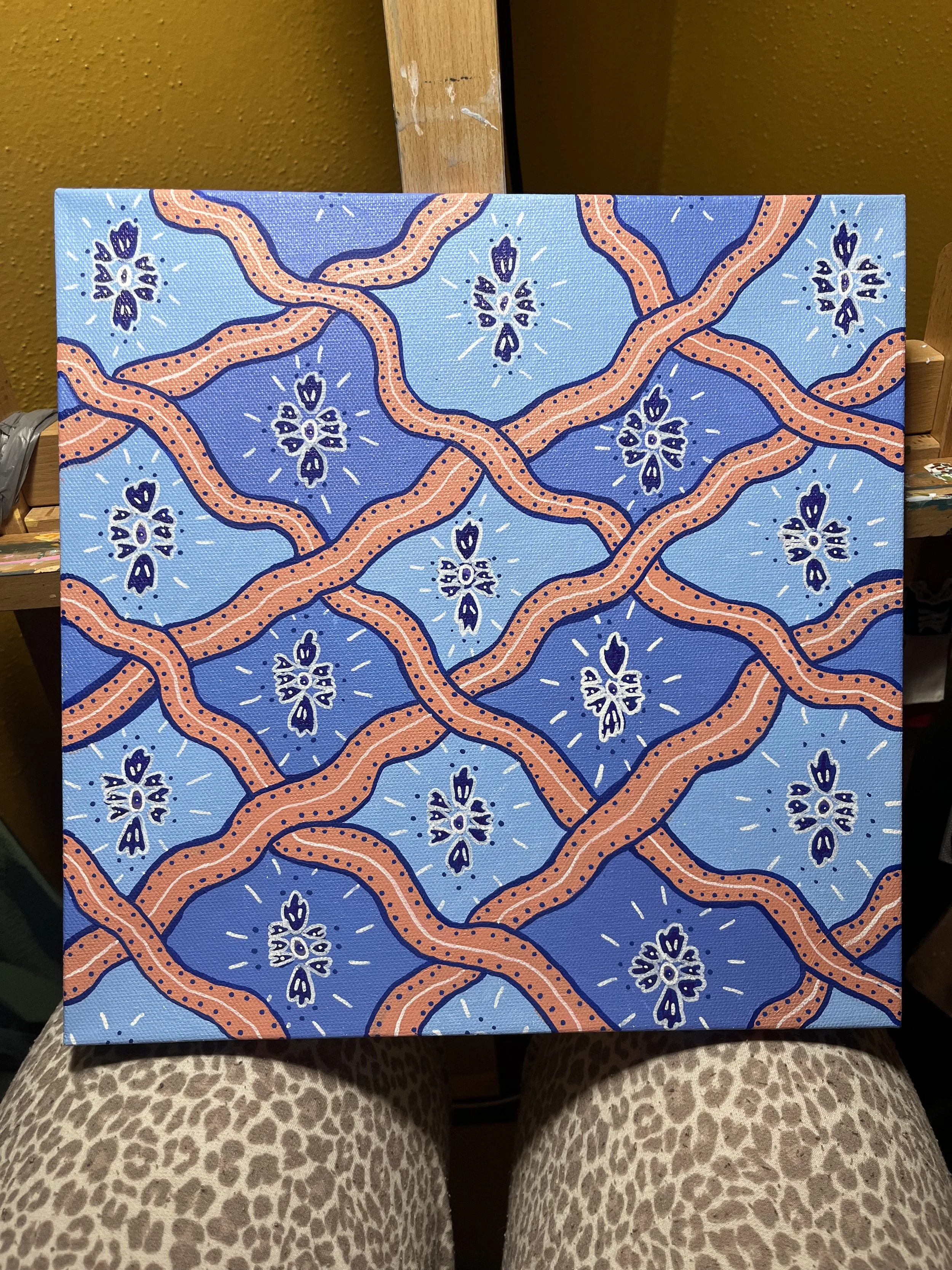 A painting with a bright blue background and orange, snake-like twisting lines. Small, symmetrical shapes resembling flowers or butterflies are scattered throughout the pattern, surrounded by white dots and lines.