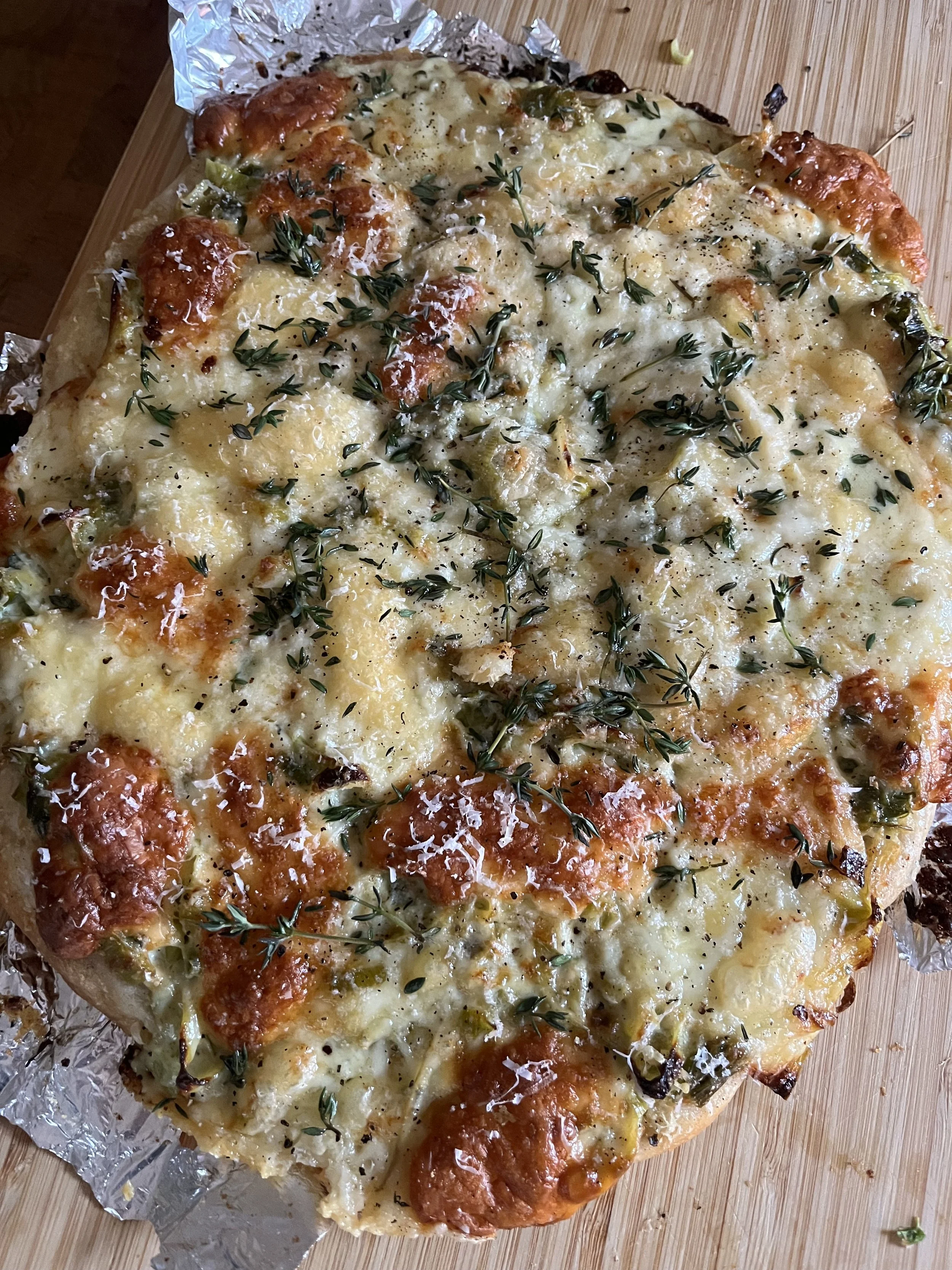 A freshly baked cheese pizza with sprigs of fresh thyme on top, served on a wooden surface with aluminum foil underneath.