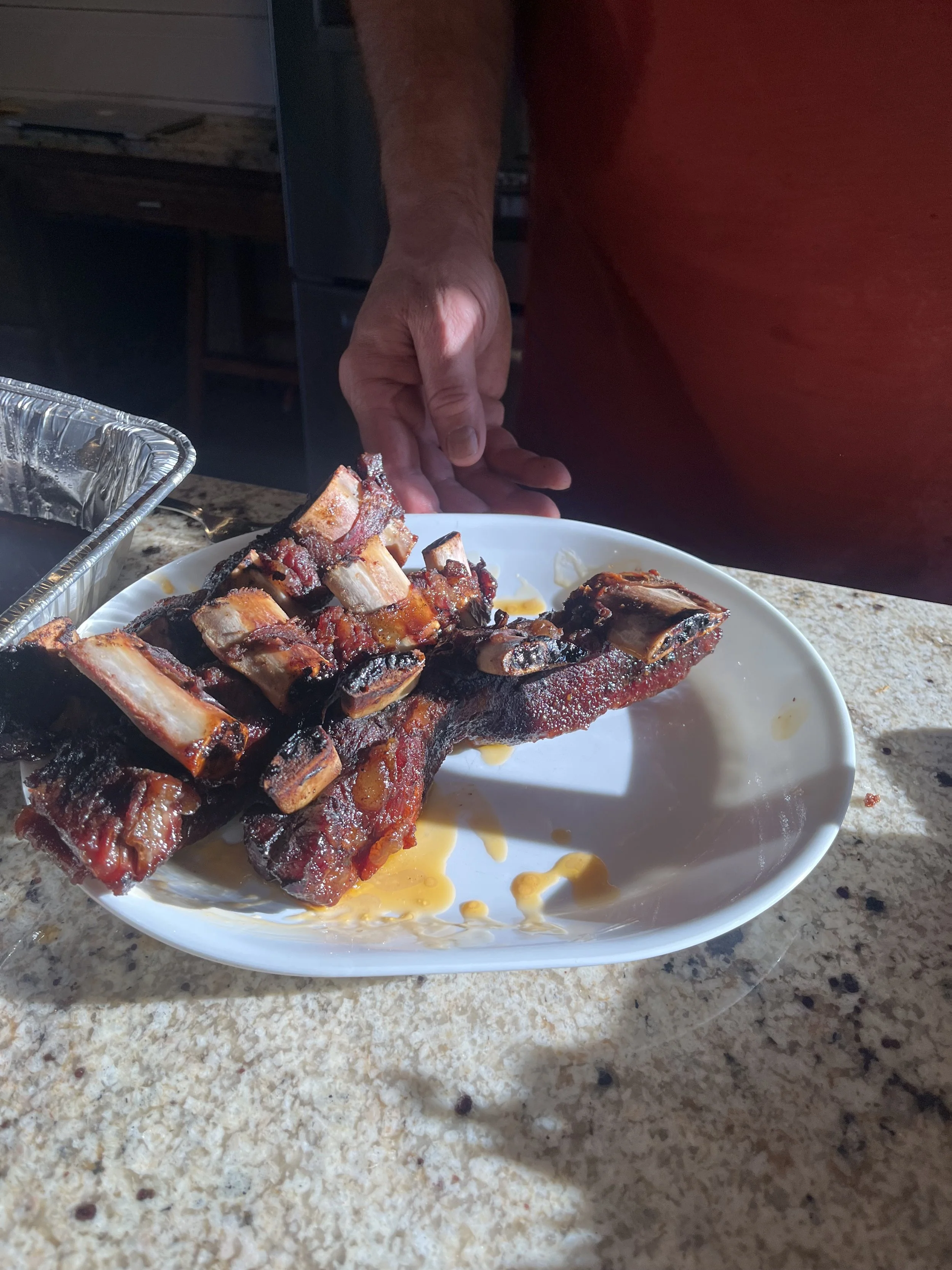 Barbecued pork ribs with a smoky glaze on a white plate, with a hand reaching for the ribs, on a granite countertop.