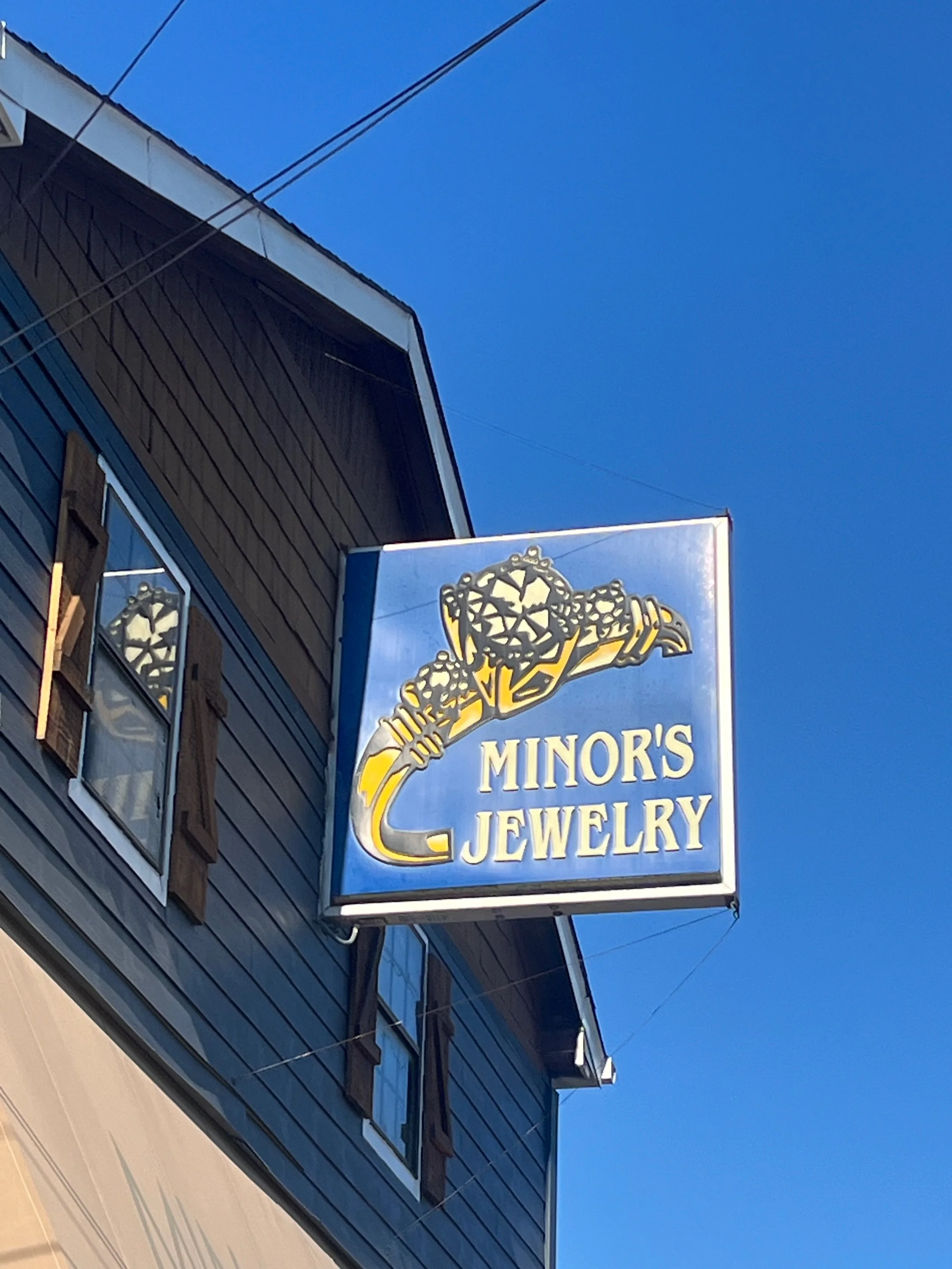 Blue sign with gold jewelry illustration and text 'Minor's Jewelry' hanging outside a building with dark blue siding and windows, under a clear blue sky.