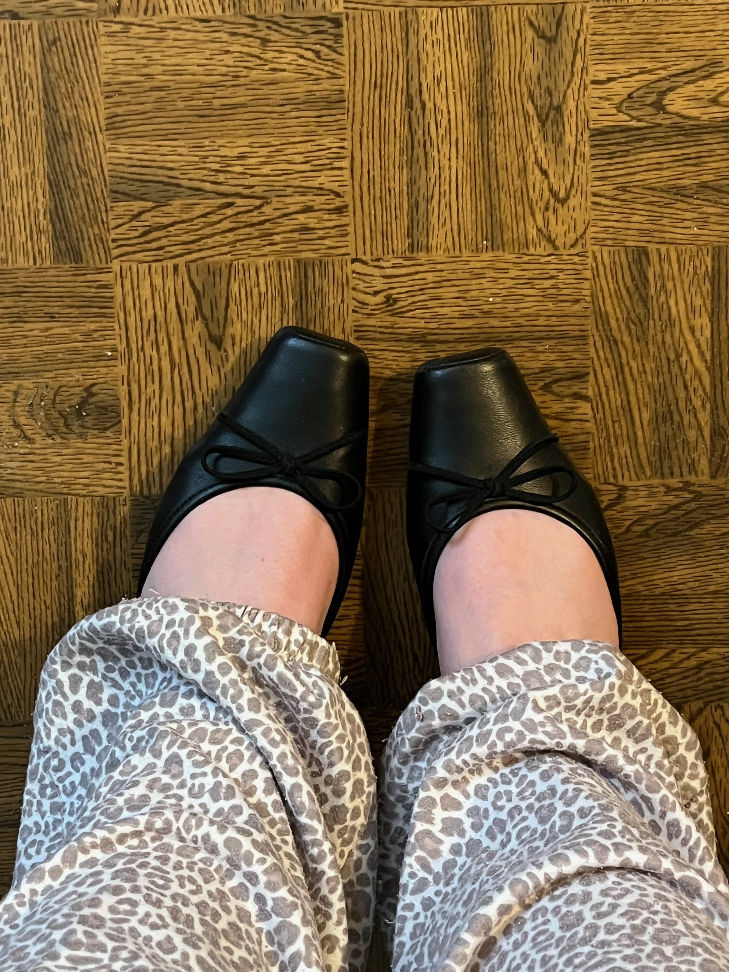 Top view of person wearing black ballet flats and leopard print pants standing on wooden floor.