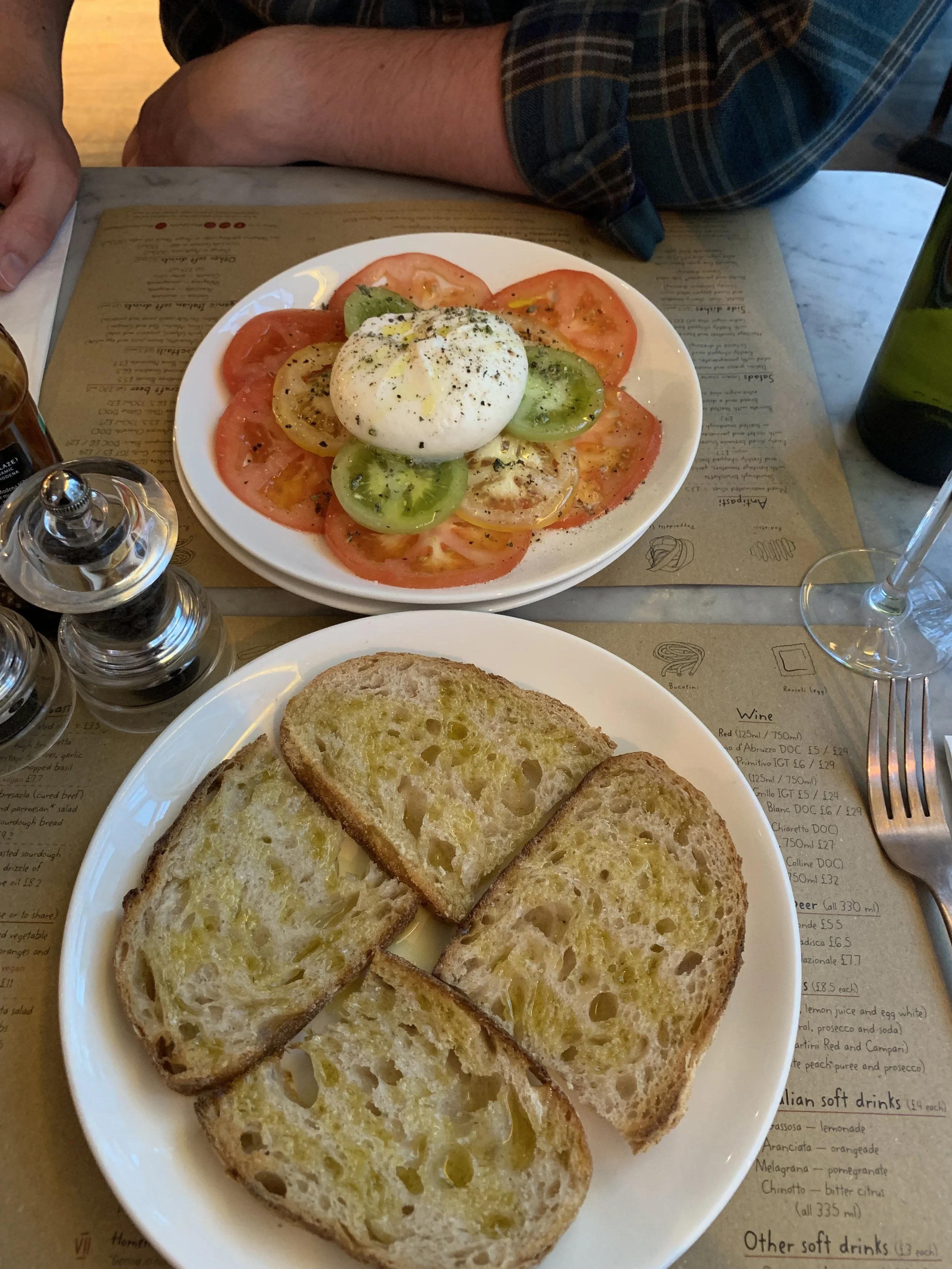 A plate of sliced toasted bread with olive oil, a bowl of sliced tomatoes, mozzarella cheese, and basil, with black pepper on top, on a table at a restaurant.