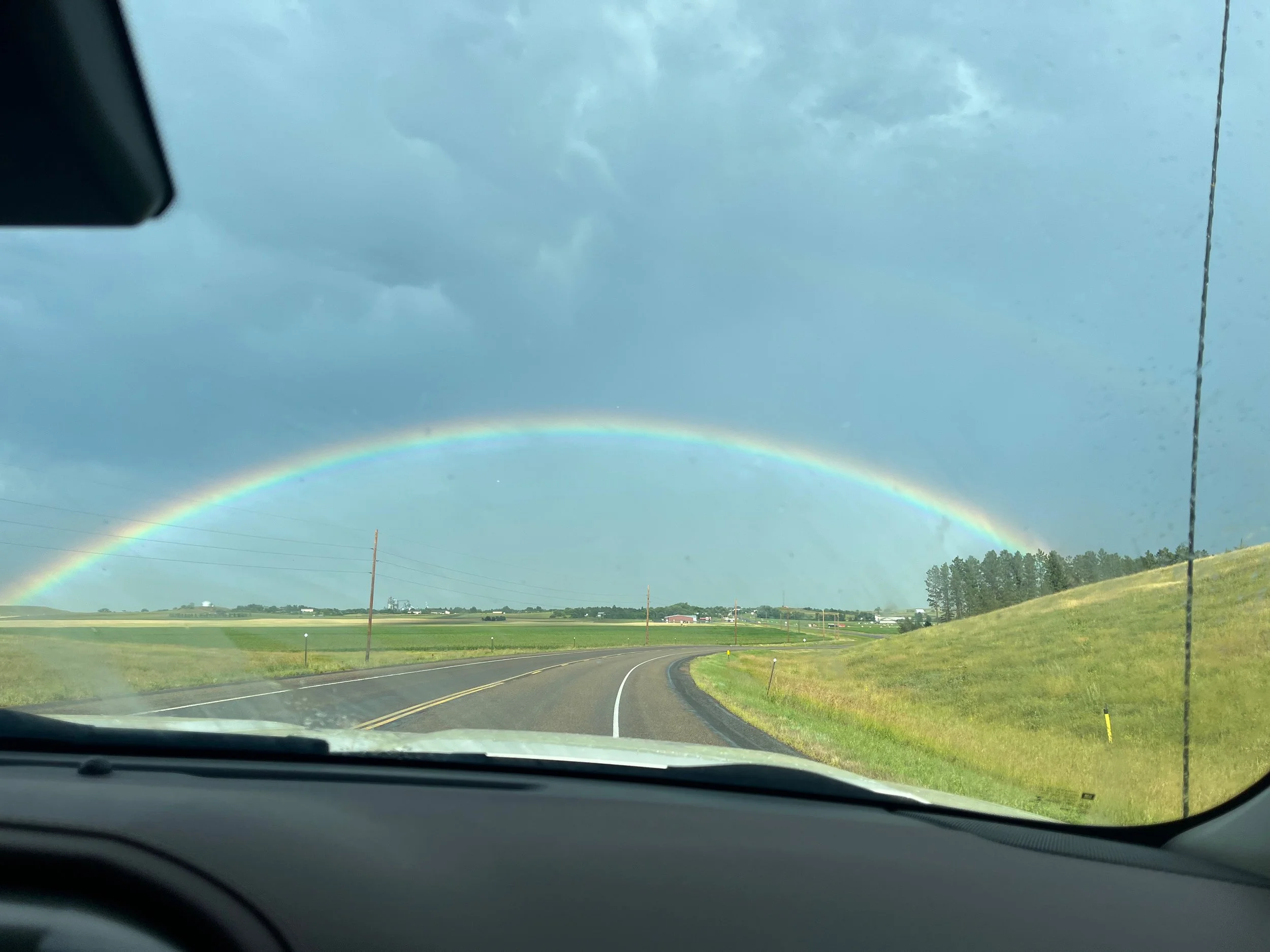 A rainbow appearing in the sky across a rural landscape viewed from inside a car, with a curving road and green fields on either side.