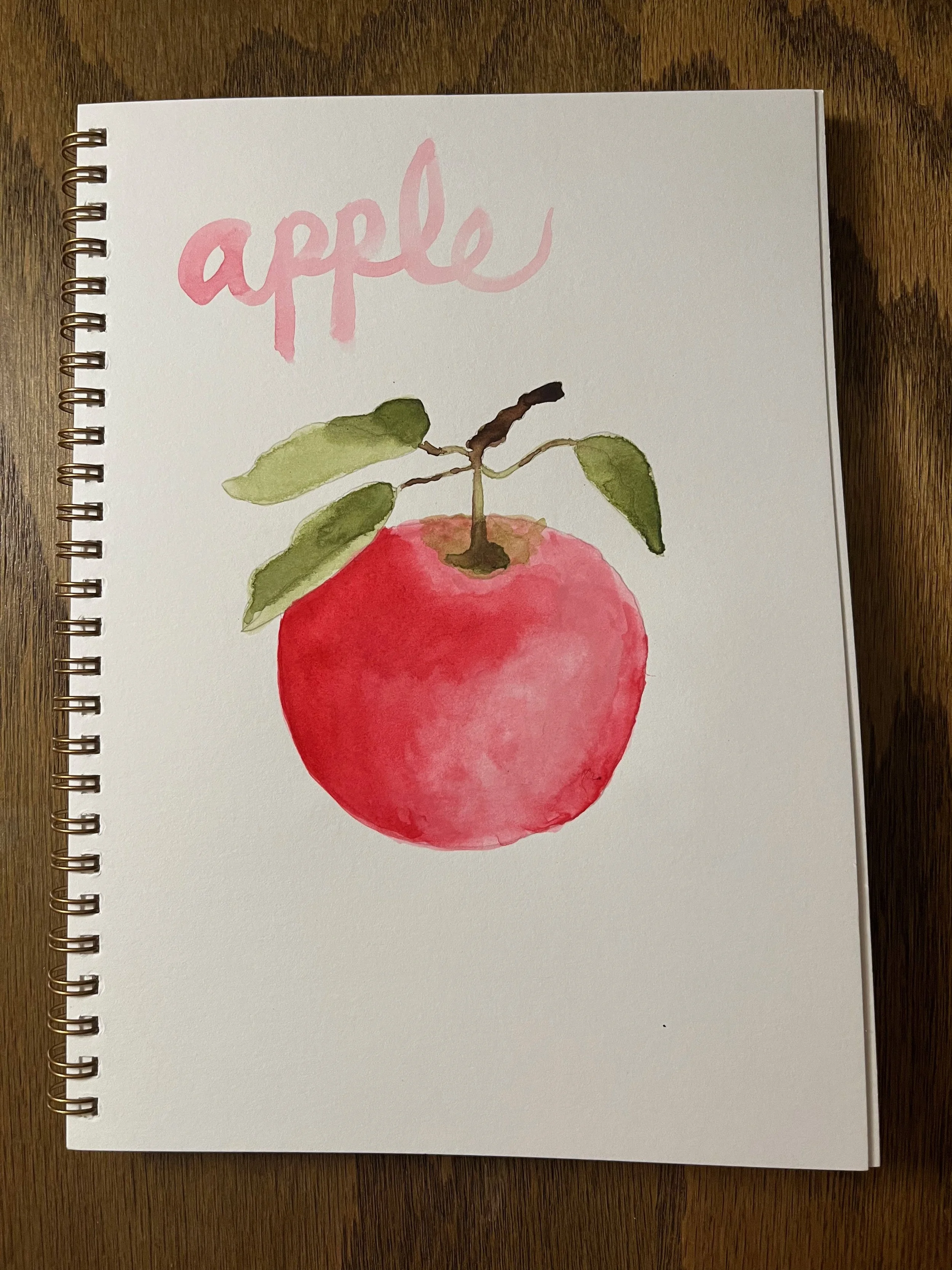 Watercolor painting of a red apple with green leaves and brown stem. The word 'apple' is written in pink above the illustration.