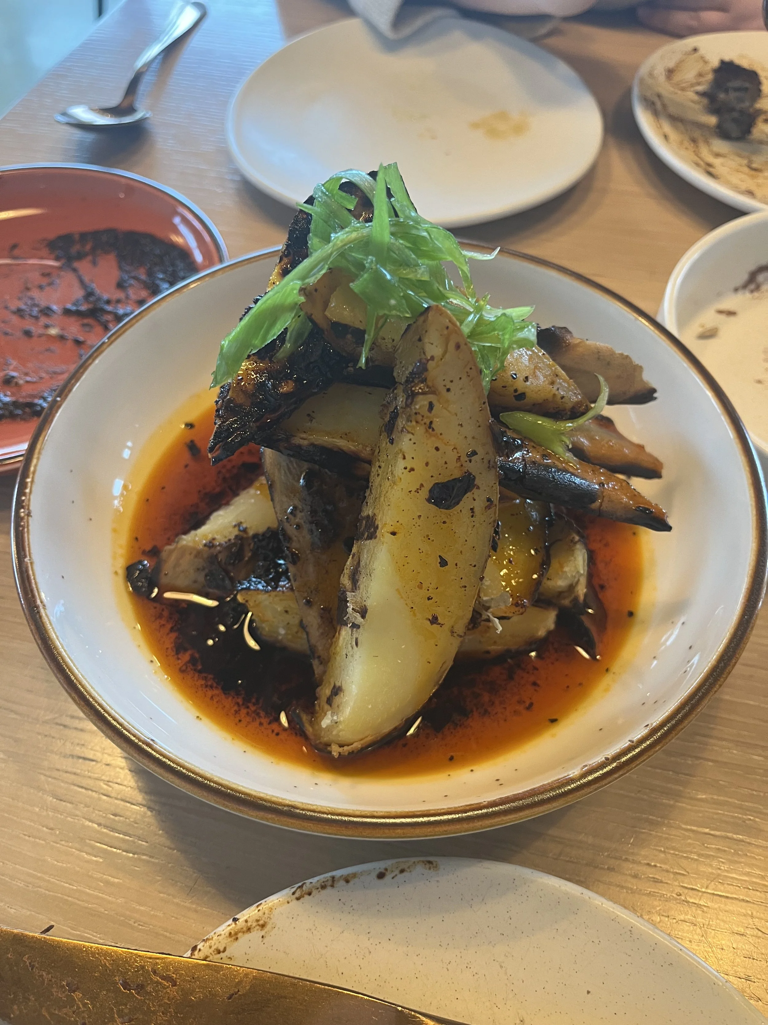 A bowl of roasted potatoes and eggplant topped with green herbs and drizzled with oil and sauce, served on a table with empty plates and utensils.