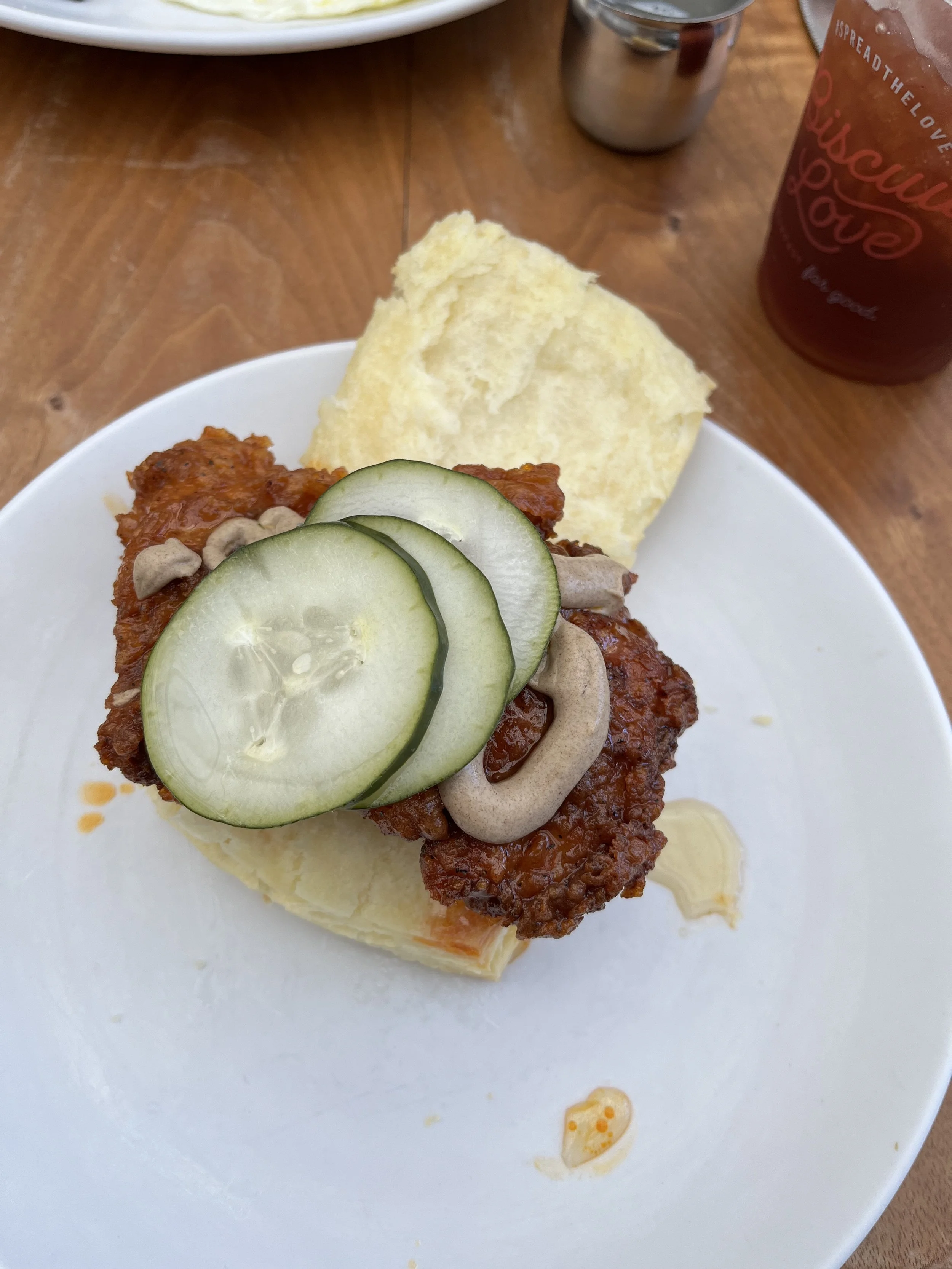 A plate with fried chicken topped with cucumber slices, some sauce, a biscuit, and some potato chips, with a glass of iced tea in the background.
