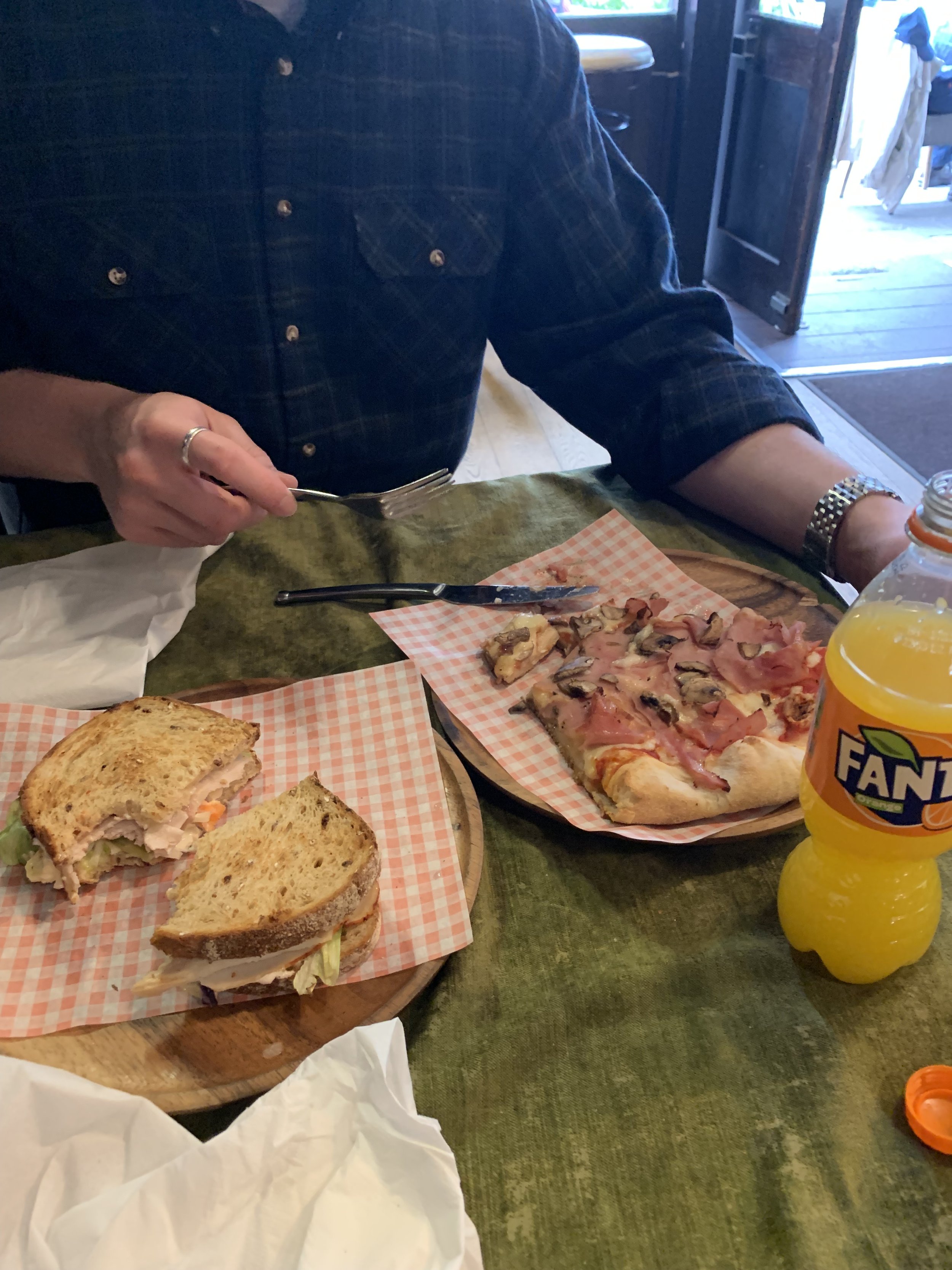 Person in a dark plaid shirt sitting at a green table, cutting a slice of pizza with toppings and a partially eaten sandwich with lettuce and meat, with a bottle of Fanta soda nearby.