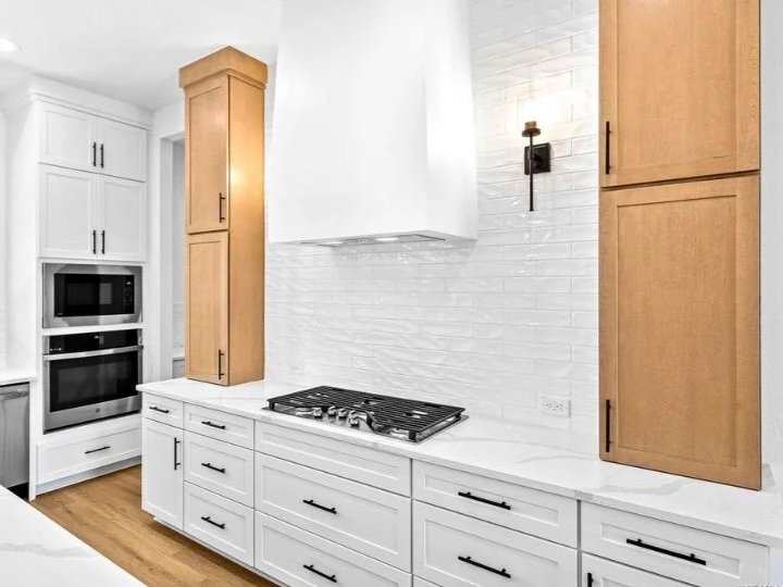 Modern kitchen with white cabinets, wood accents, a built-in microwave and oven, and a gas stovetop. White brick backsplash and a black wall sconce light.