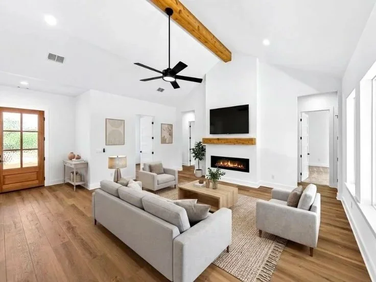 Bright living room with white walls, wooden floor, modern furniture, a fireplace, a large flat-screen TV, ceiling fan, and large windows.
