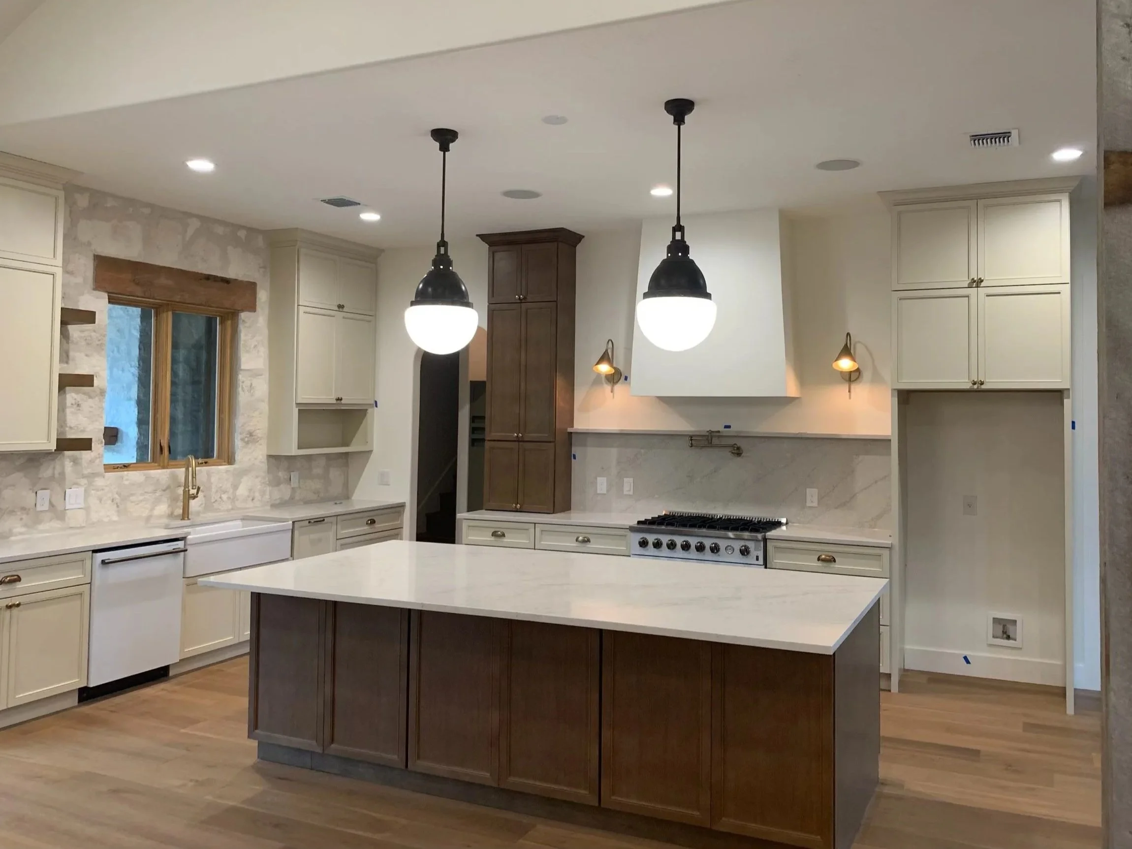 A modern kitchen with white cabinets, a large central island with a wooden base, a white marble countertop, pendant lights, and a window above the sink.