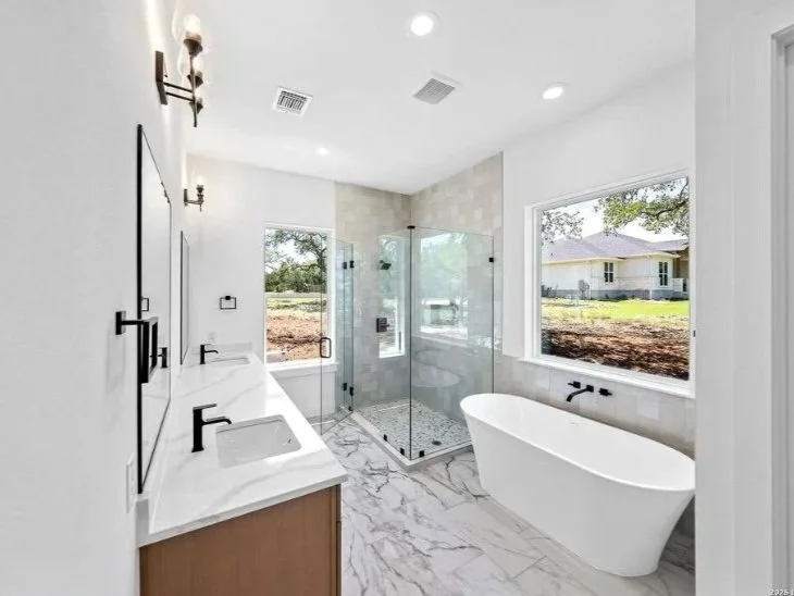 Modern bathroom with a double vanity, a freestanding bathtub, and a glass-enclosed shower, illuminated by natural light from two large windows.