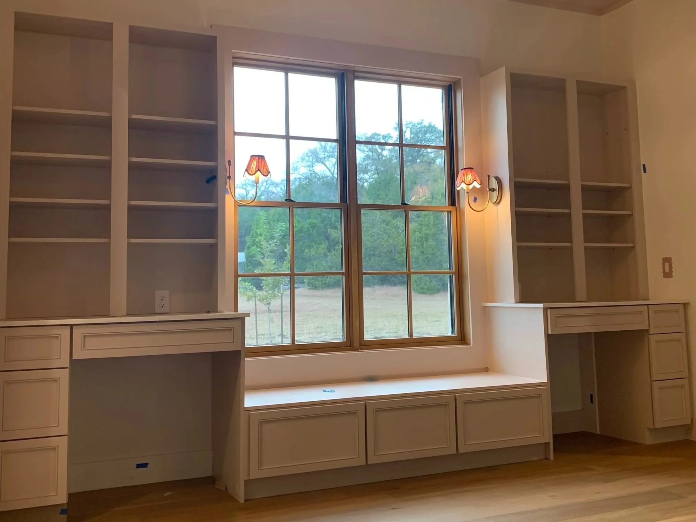 Empty built-in bookshelves and window seat in a room with two wall sconces, large window, and hardwood floors.