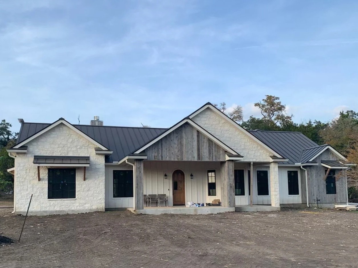Newly constructed house with a black metal roof, white stone, and wood siding, on a dirt lot with trees in the background.