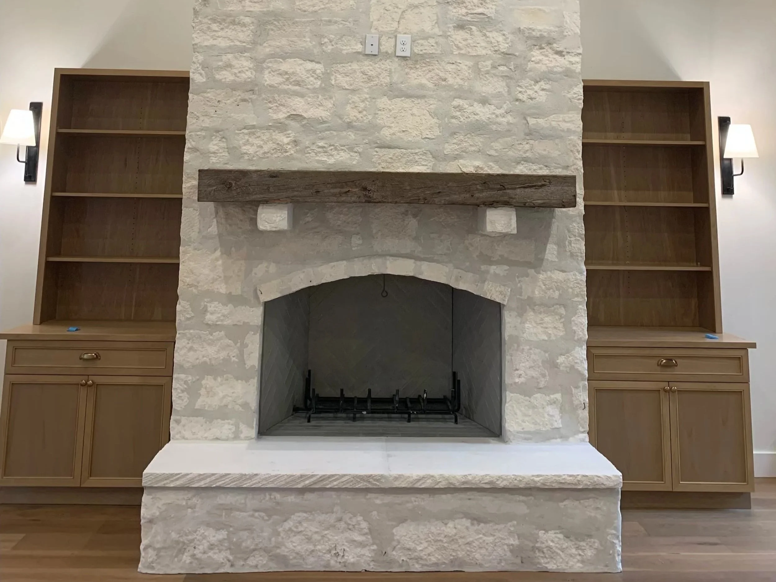 Living room fireplace with white stone surround, wooden mantle, and built-in bookshelves on each side, flanked by wall-mounted lamps.