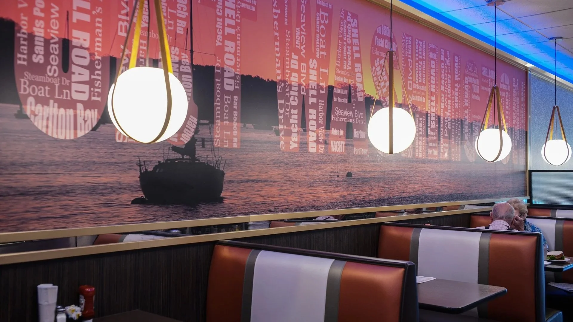 A restaurant interior with booth seating and a large wall mural depicting a boat on water at sunset. The mural has a collage of pink and purple street names related to Yachting and waterfront areas. There are four hanging pendant lights with white gl