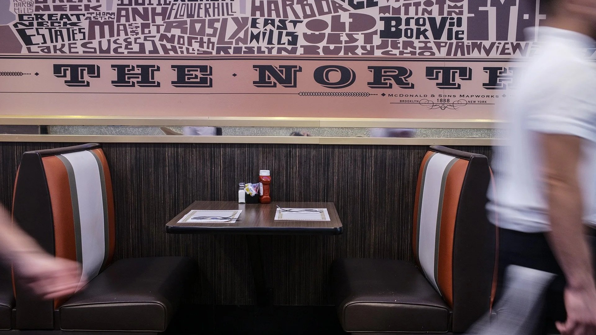 Empty booth at a restaurant with two placemats, ketchup and mustard bottles on the table, and two chairs with brown, black, and white upholstery. Partially visible people are walking past, and a large wall decoration with text reading 'The North' is 