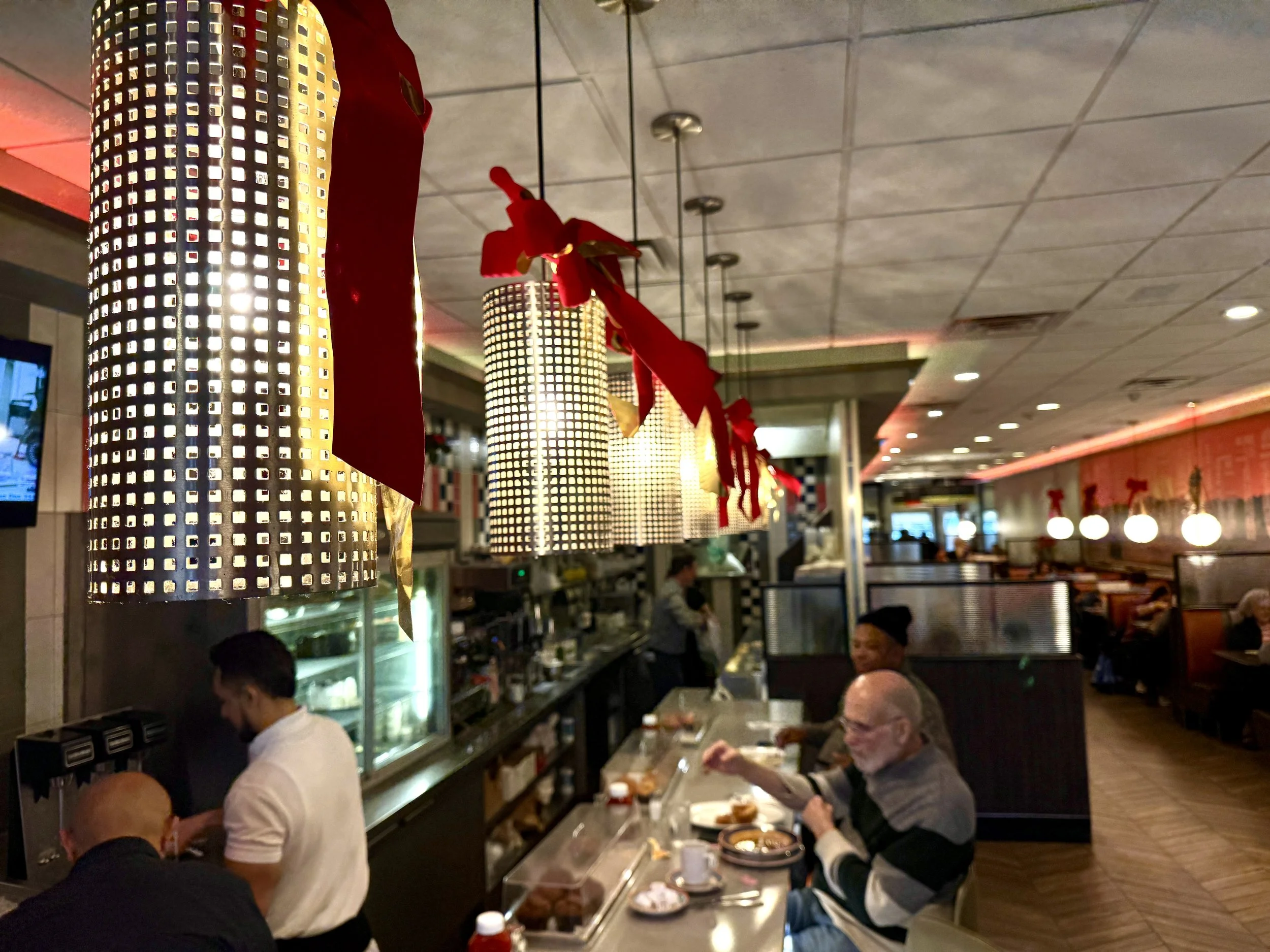 Interior of a restaurant decorated for Christmas with hanging lights resembling giant lanterns with red ribbons, booths and tables with people dining, and a counter with a staff member serving food.