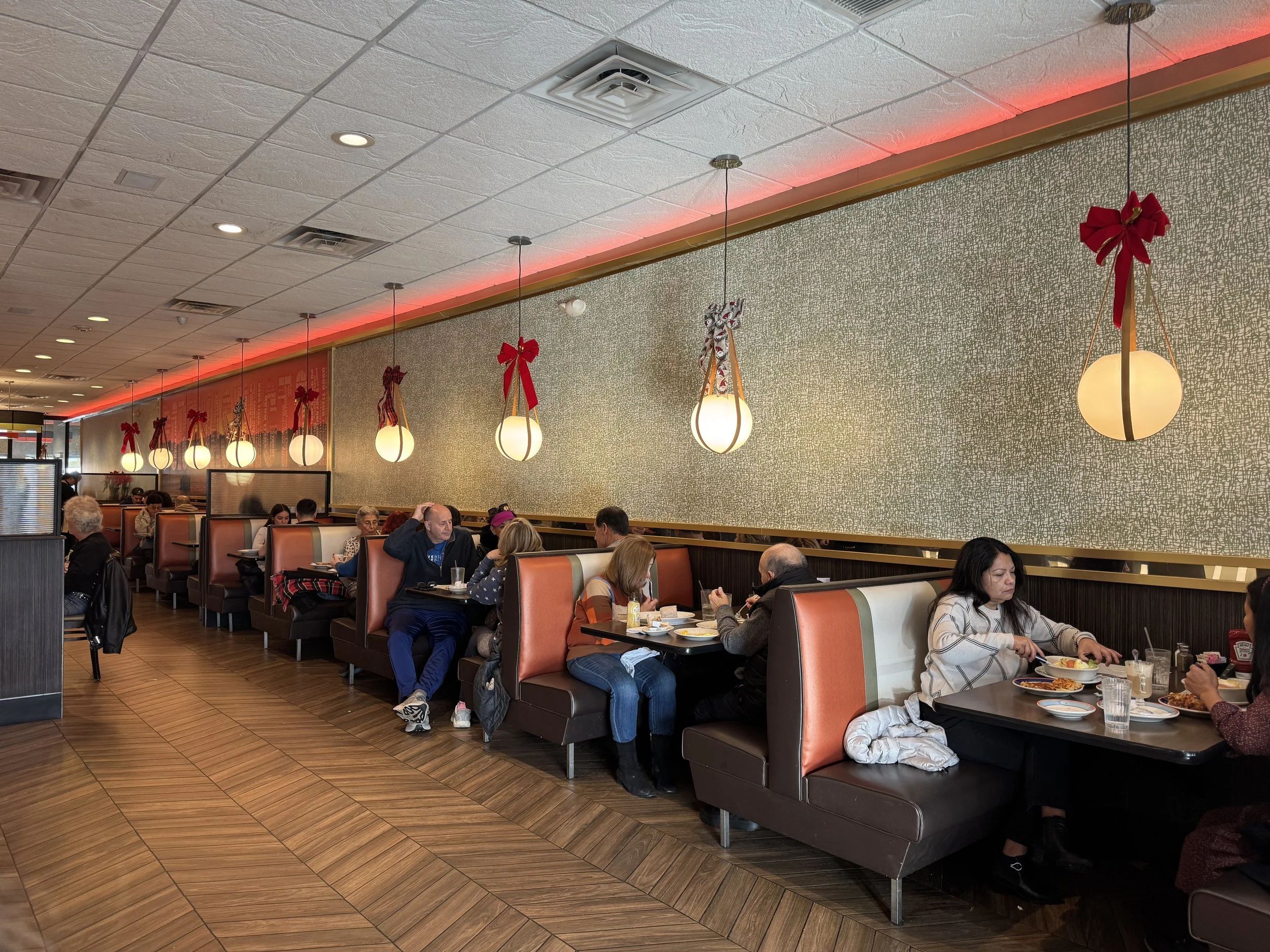 A busy restaurant interior decorated with Christmas ornaments, including hanging lights with red bows and ribbons. Customers sit in booths eating and drinking, with wood flooring and textured wallpaper behind the booths.