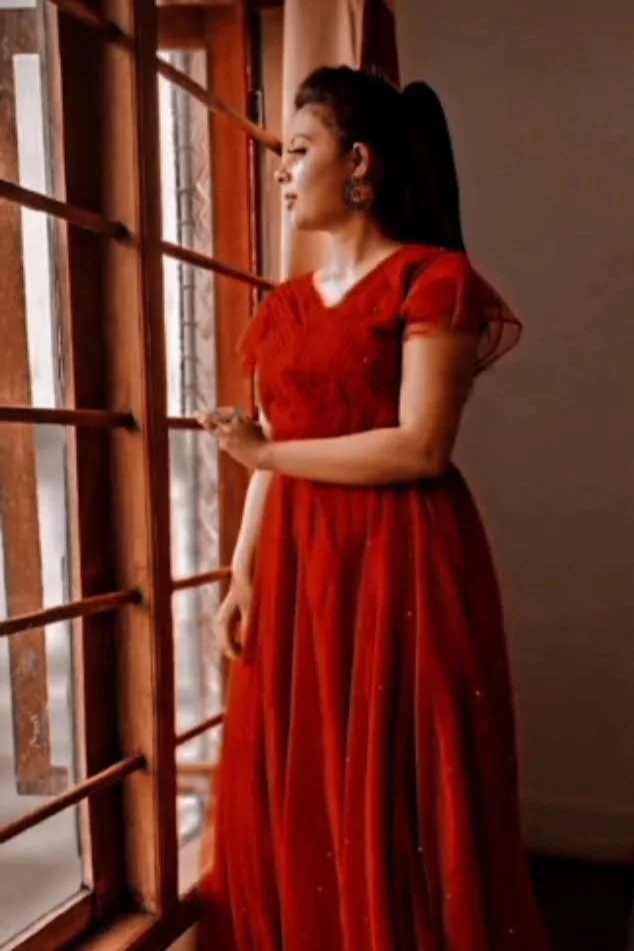 Woman in red dress standing by window, looking outside.