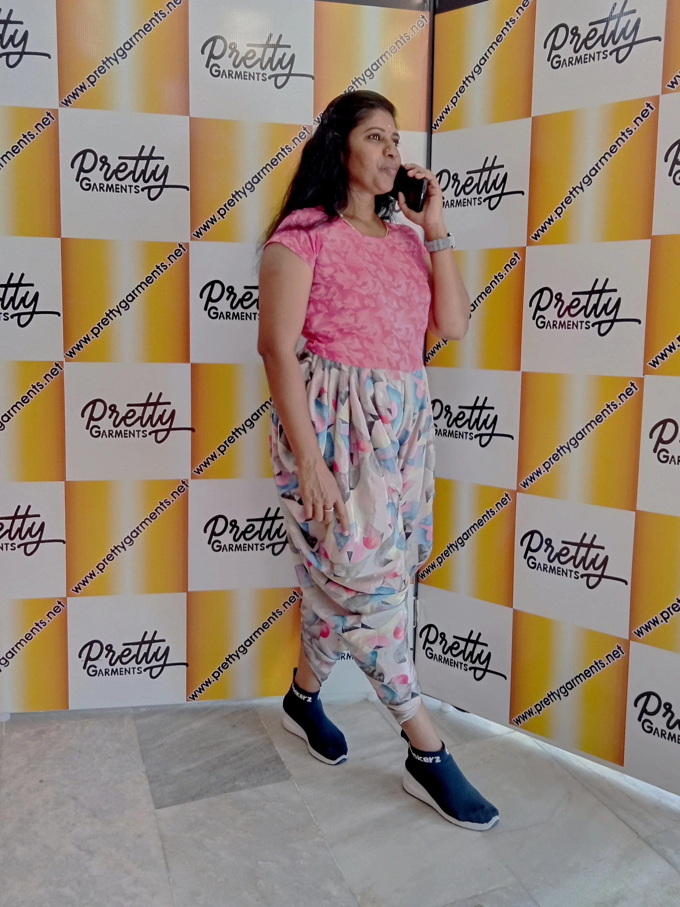 Woman in colorful outfit standing in front of a photo wall with "Pretty Garments" branding, talking on a smartphone.