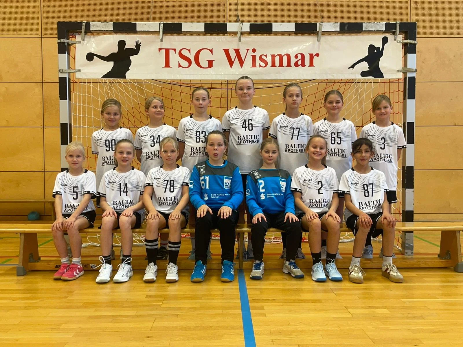 Mädchen-Handballmannschaft in roter Teamkleidung auf einem Hallenboden, mit einem Tor im Hintergrund und zwei Spielerinnen in grüner Kleidung liegend vorne.