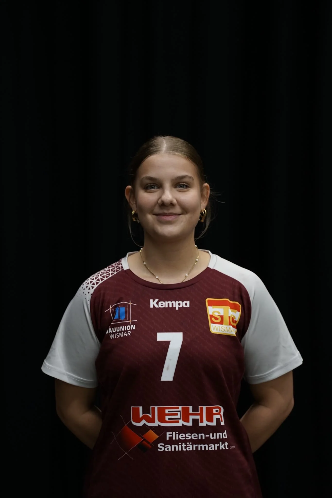 Junge Frau in einem weißen Sporttrikot mit der Nummer 77 und Logos auf der Vorderseite steht vor einem grünen Netz.