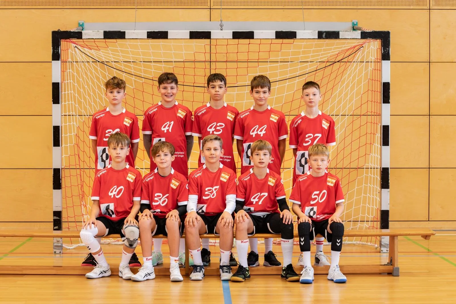 Eine Gruppe von jugendlichen Handballspielern posiert lächelnd für ein Foto in einer Sporthalle. Einige tragen rote und weiße Sporttrikots, ein Erwachsener ist als Weihnachtsmann verkleidet. Ein Fußballtor ist im Hintergrund sichtbar.