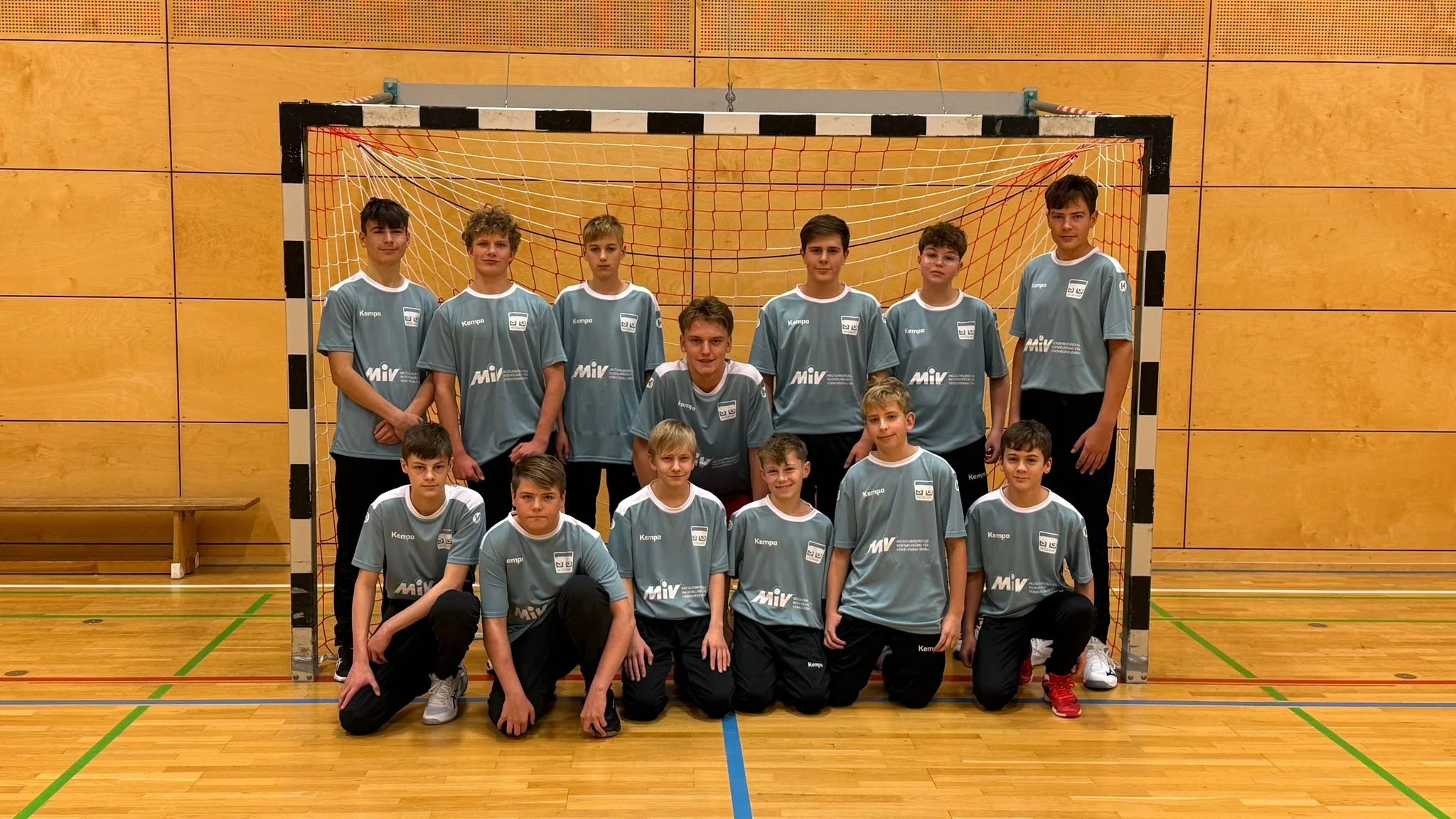 Handballteam mit Spielern und Trainern in einer Sporthalle vor einem Tor posierend, alle in Trikots.