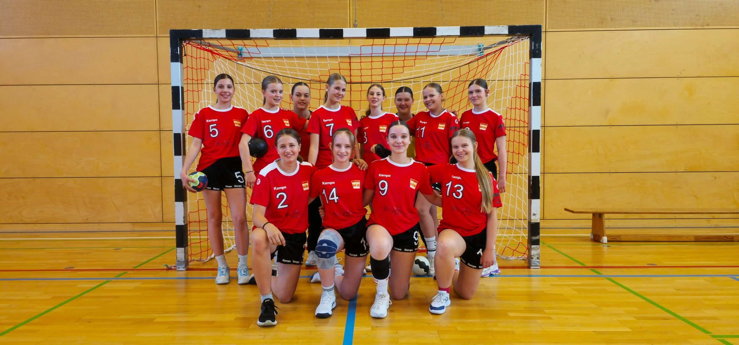 Eine Gruppe von Mädchen in Handballtrikots posiert vor einem Handballtor in einer Sporthalle. Die meisten tragen Medaillen um den Hals, und einige haben Weihnachtsmützen auf. Sie lächeln und scheinen ein Team zu sein, das an einem Turnier teilgenommen hat.