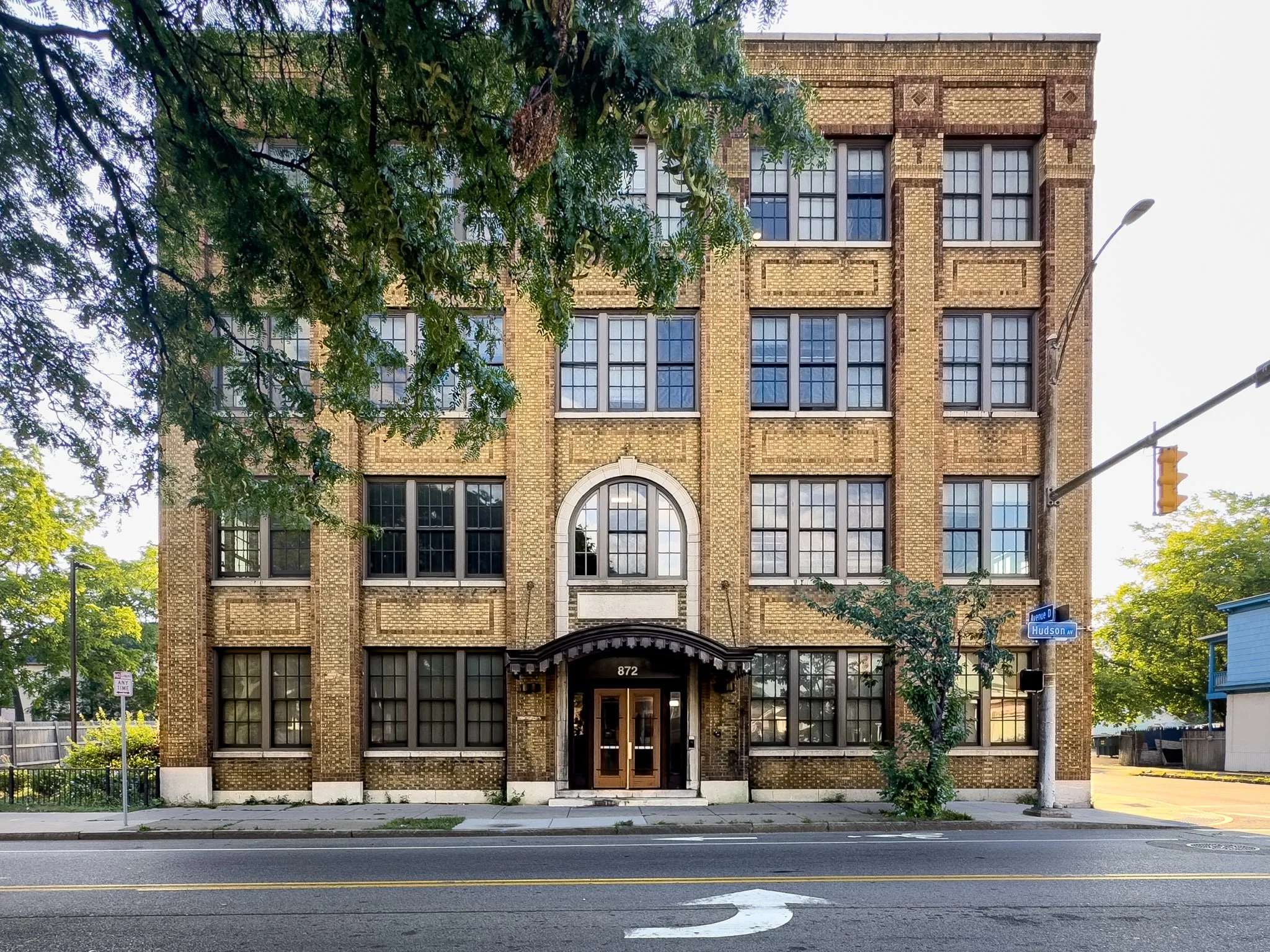 A multi-story brick building with large grid-patterned windows and an arched entrance at the front, situated on a city street with a sidewalk, street corner, and traffic light, surrounded by green trees.