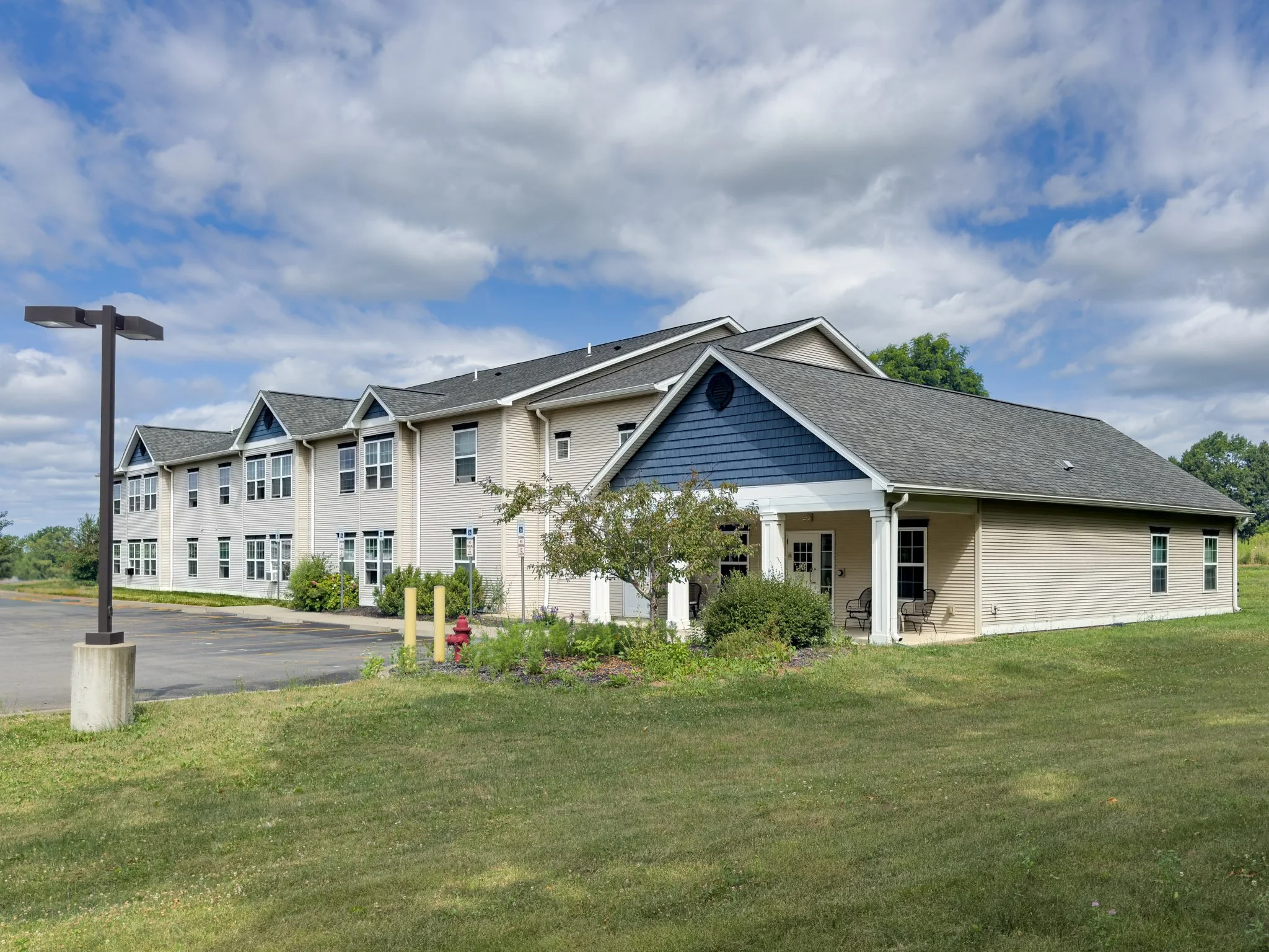 A multi-story residential building with beige siding and blue accents under a partly cloudy sky. There is a parking lot with a street lamp, a fire hydrant, and some bushes in the foreground.