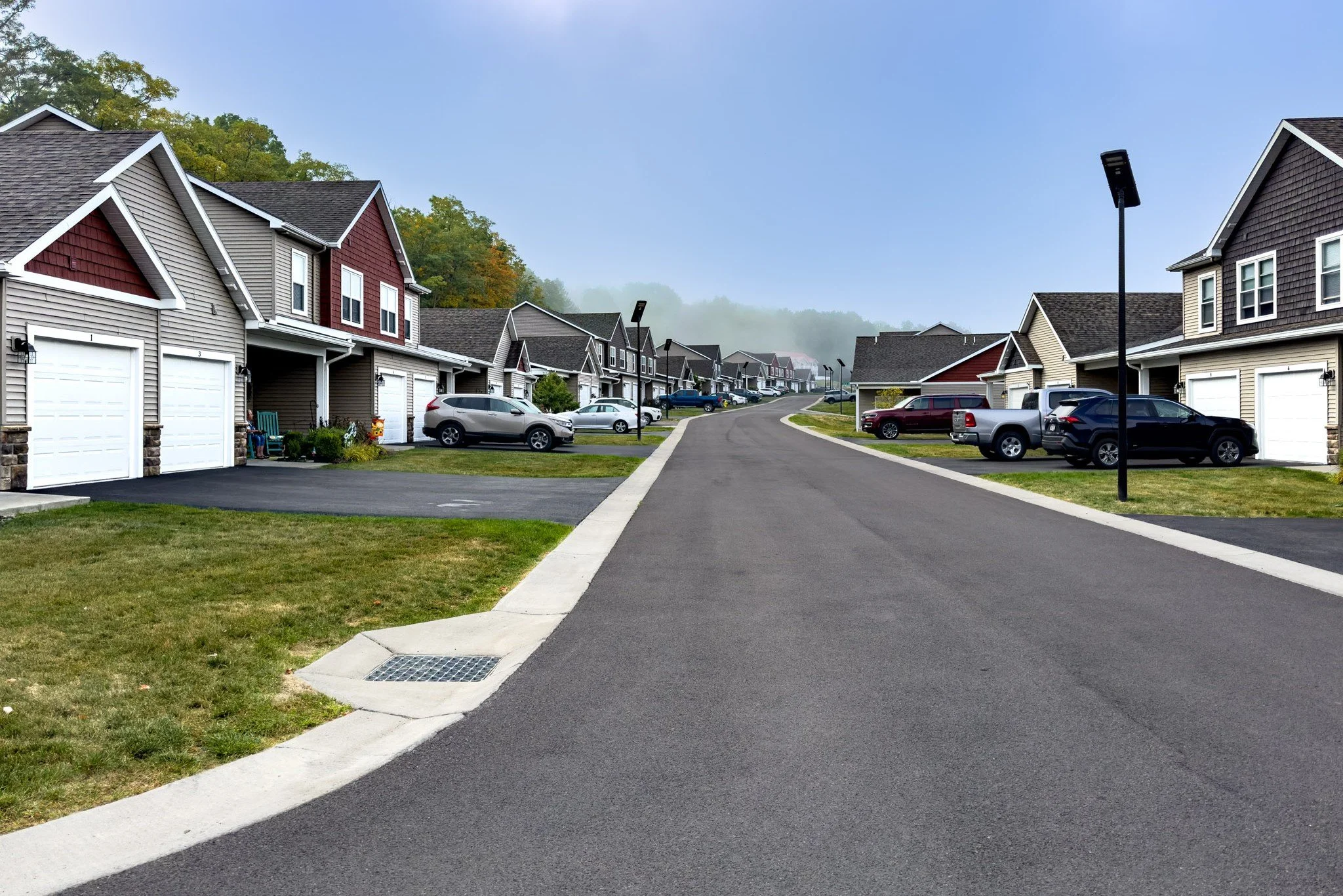 A suburban street with single-family homes, parked cars, street lamps, and a grassy strip along the sidewalk, with a hill and trees in the background.