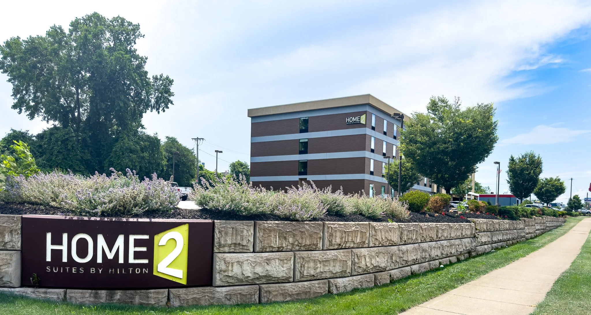 Exterior view of a modern, multi-story hotel building with the sign 'HOME2 Suites by Hilton' on a stone wall in the foreground, surrounded by trees and landscaped bushes under a partly cloudy sky.