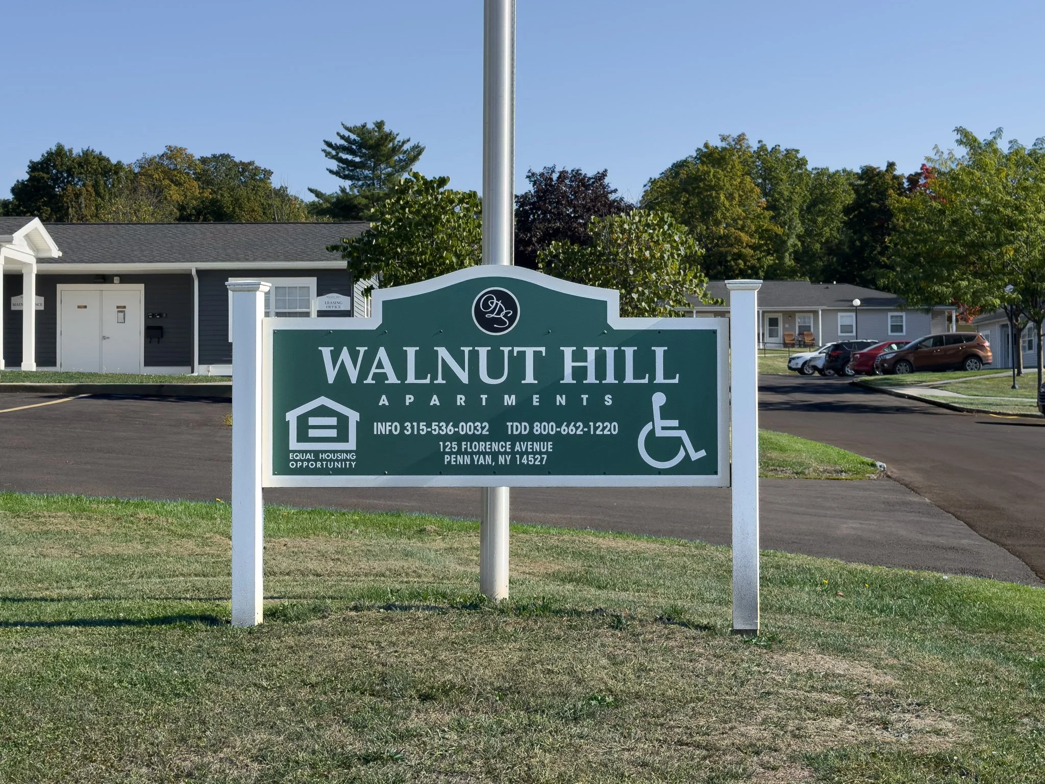 Sign for Walnut Hill Apartments with contact information and accessibility symbols, situated in a landscaped area with trees and buildings in the background under a clear blue sky.