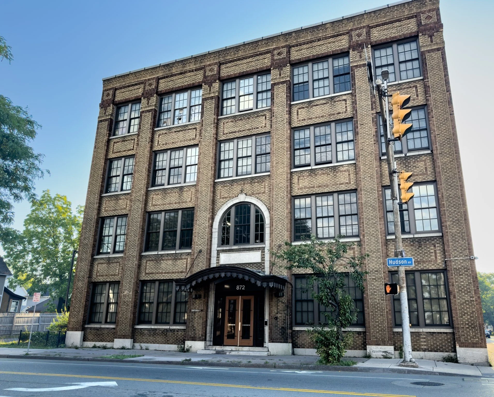 A multi-story brick building with large grid windows and an arched entrance, situated on a city street corner. A traffic light and street sign reading "Hudson Av" are visible in the foreground, with trees and residential houses in the background.