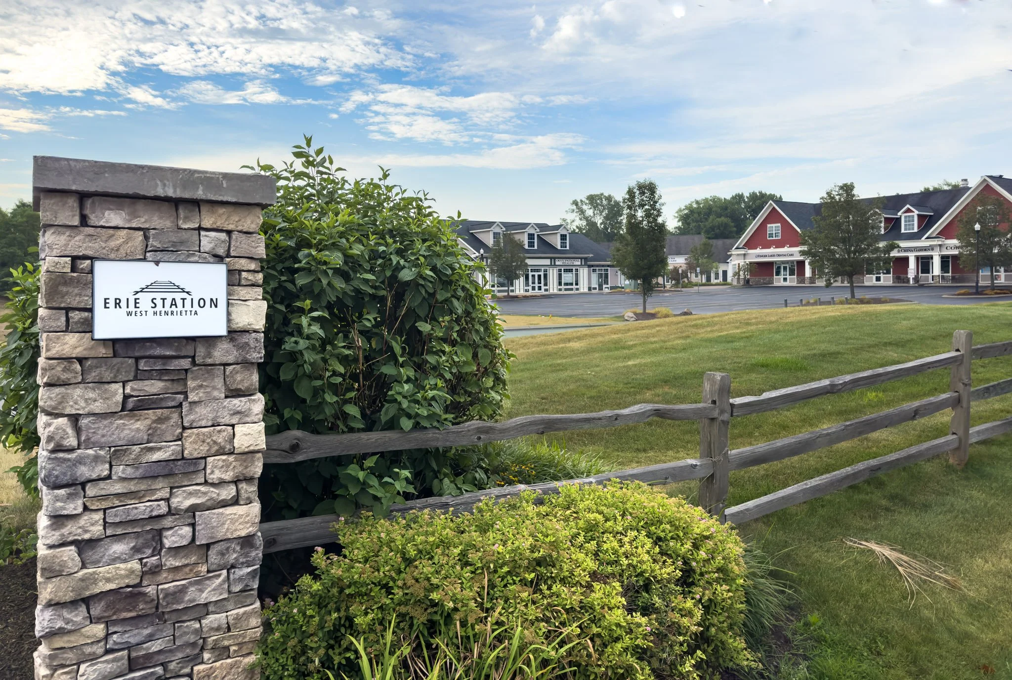 Sign in front of a shopping center reads ERIE STATION WEST HENRIETTA. There is a stone post, green bushes, and a wooden fence in the foreground. In the background, there are several storefronts, a parking lot, trees, and a partly cloudy sky.