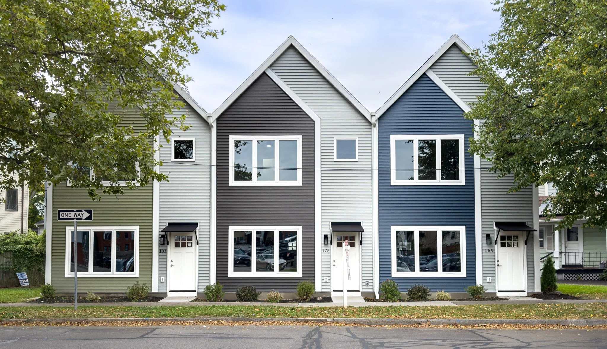 Colorful row of townhouses with different shades of blue, green, and beige, with trees partially covering the top and a one-way street sign at the corner.