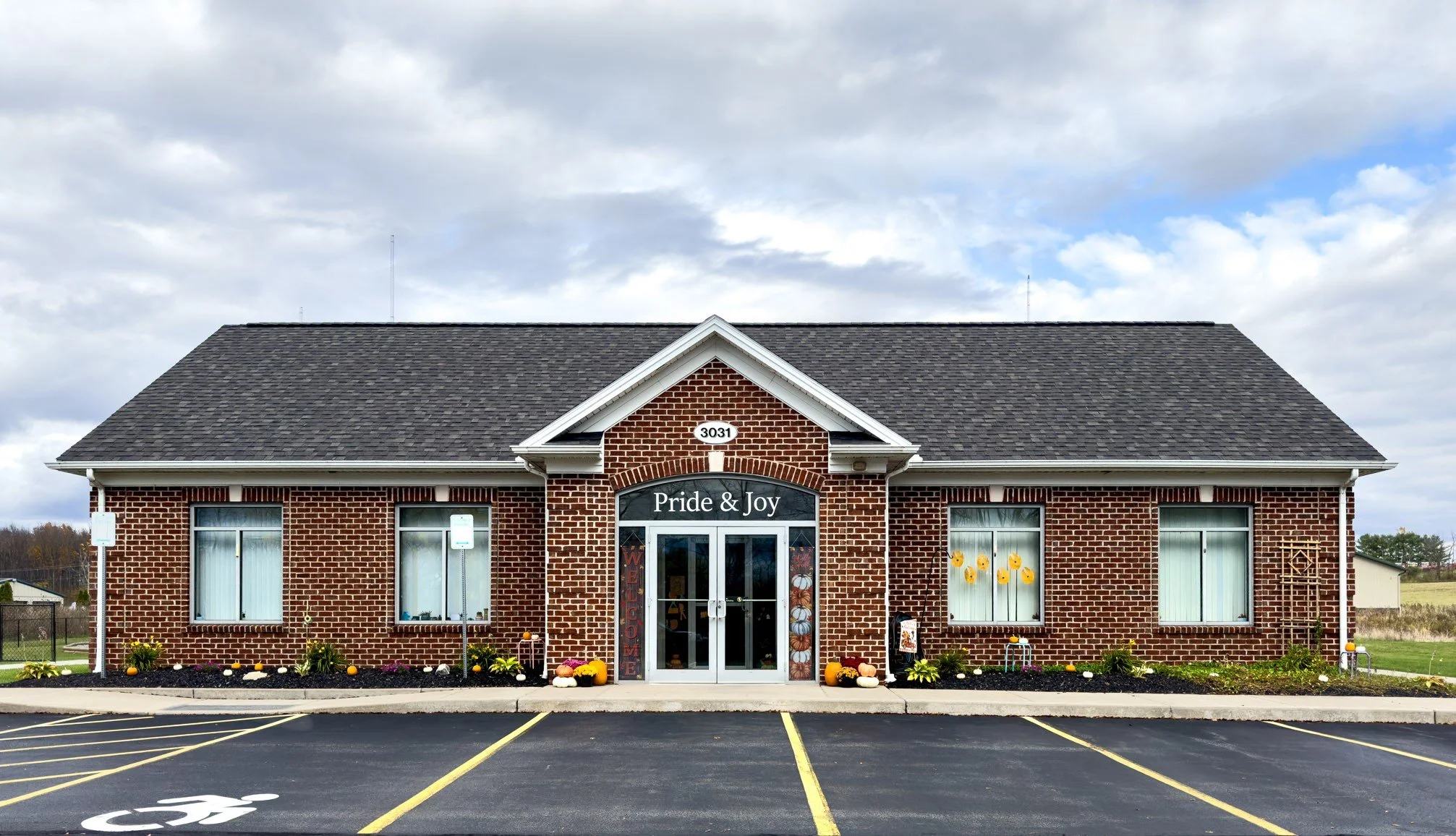 A brick building with the sign 'Pride & Joy' above the entrance, decorated with fall-themed flowers, pumpkins, and scarecrows, and surrounded by empty parking spaces in a lot.
