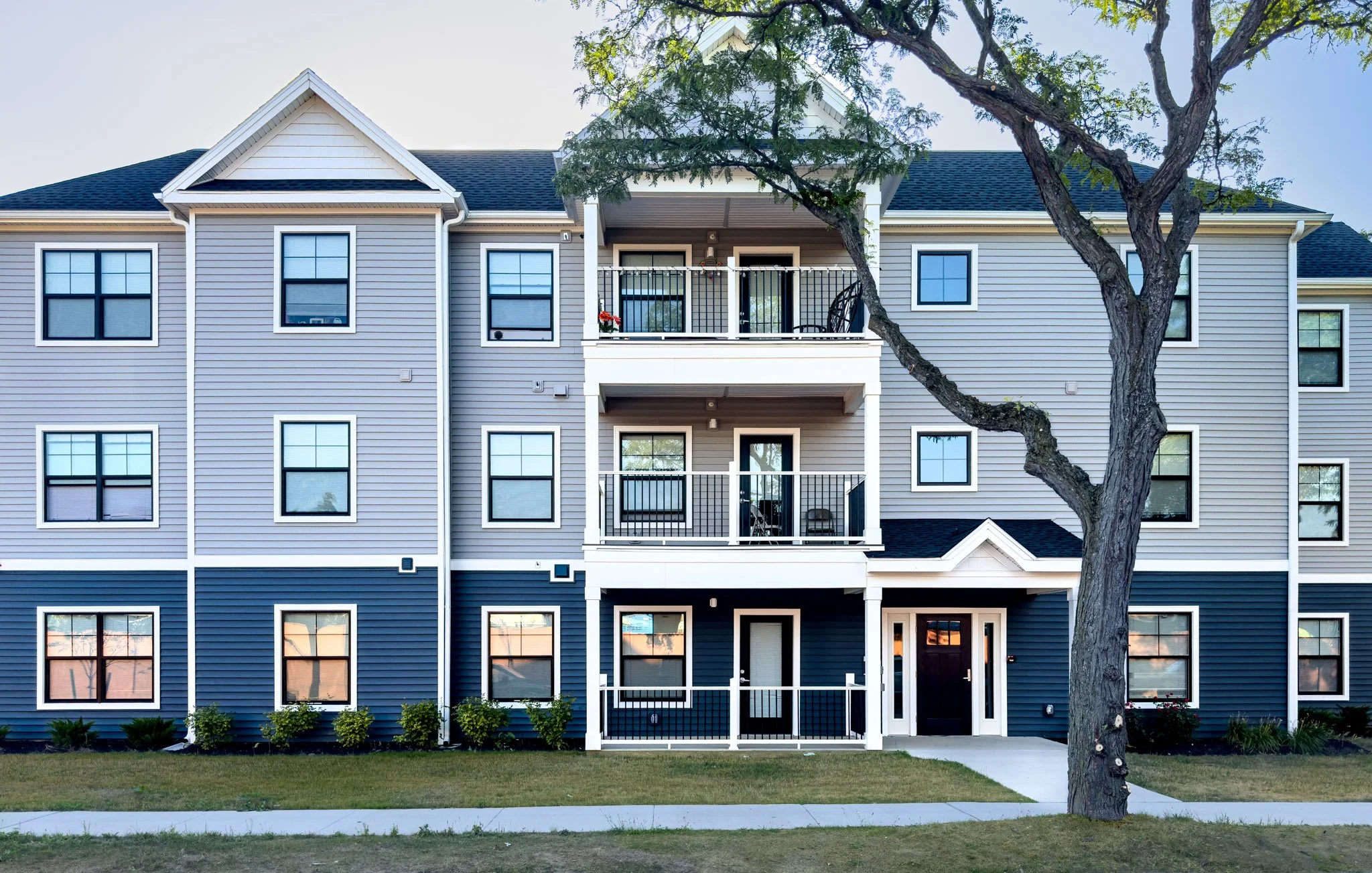 Front view of a multi-story residential apartment building with blue siding, white trim, and black window frames. There is a sidewalk and a grassy lawn with small bushes and a large tree in the foreground.