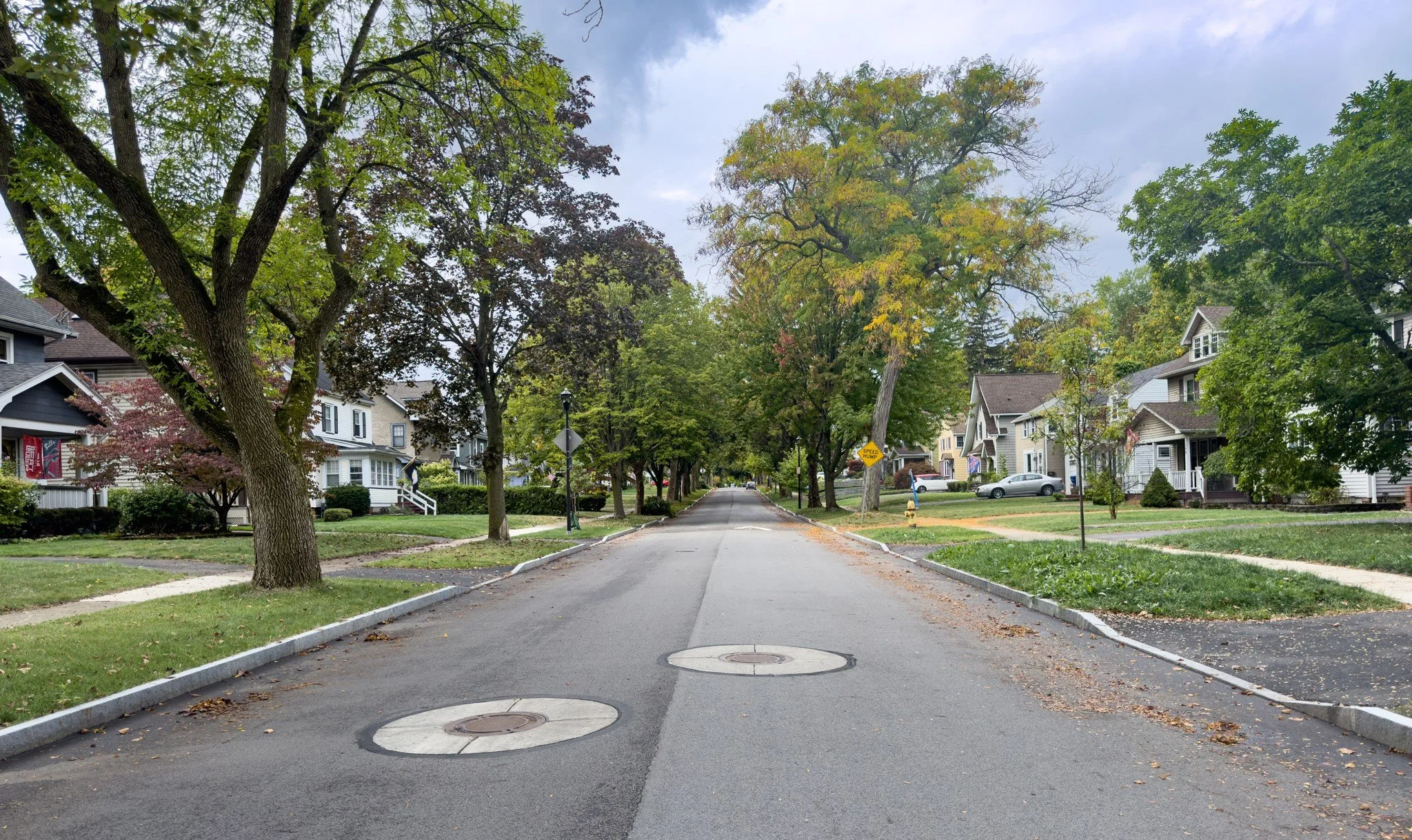 A residential street lined with trees and houses on both sides, with some trees showing early signs of changing leaves, indicating fall.