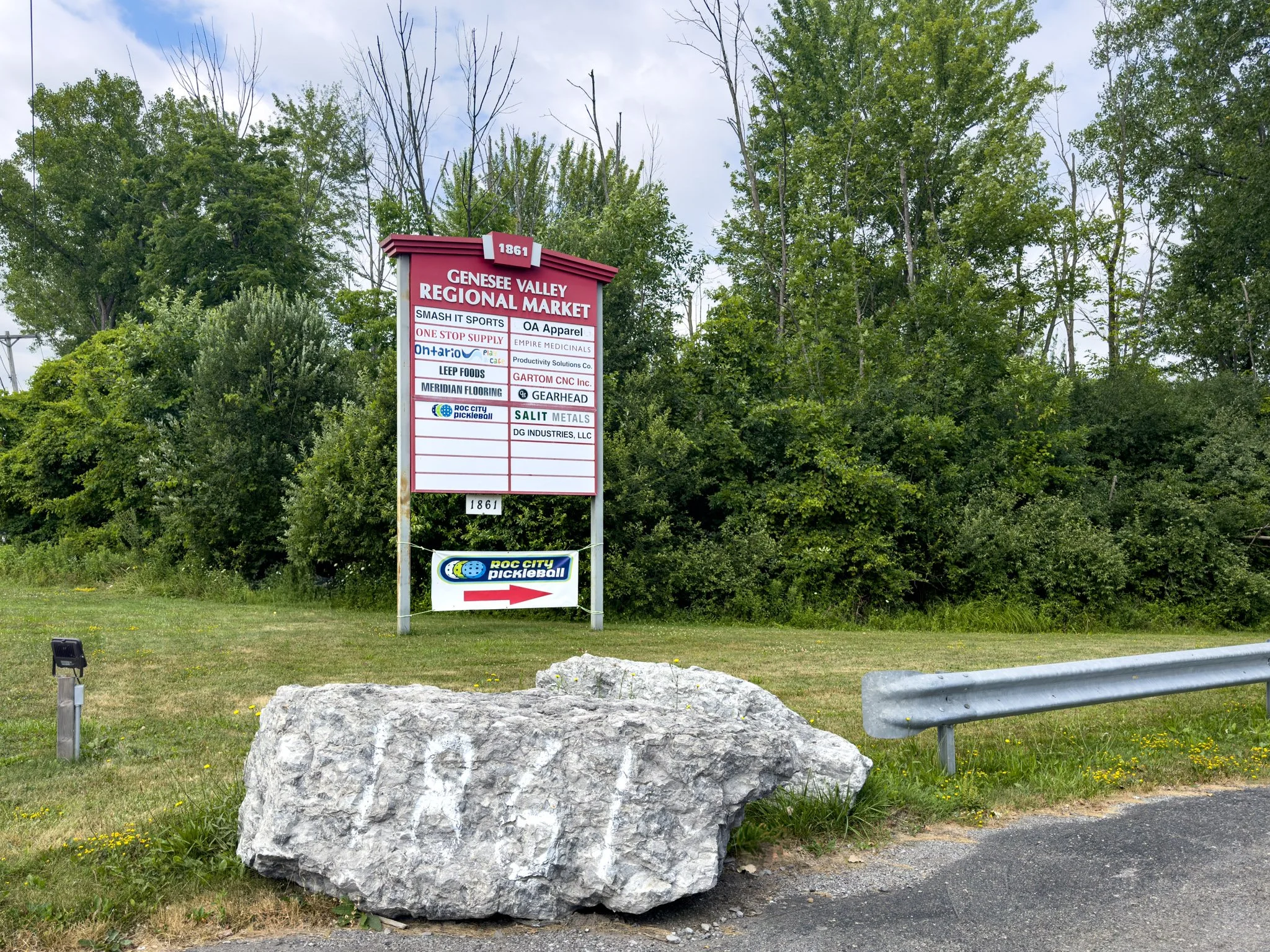 A large sign for the Genesee Valley Regional Market standing next to a row of trees and a grassy roadside, with a smaller arrow sign for Roc City Pickleball in front, near a guardrail and a large white rock.