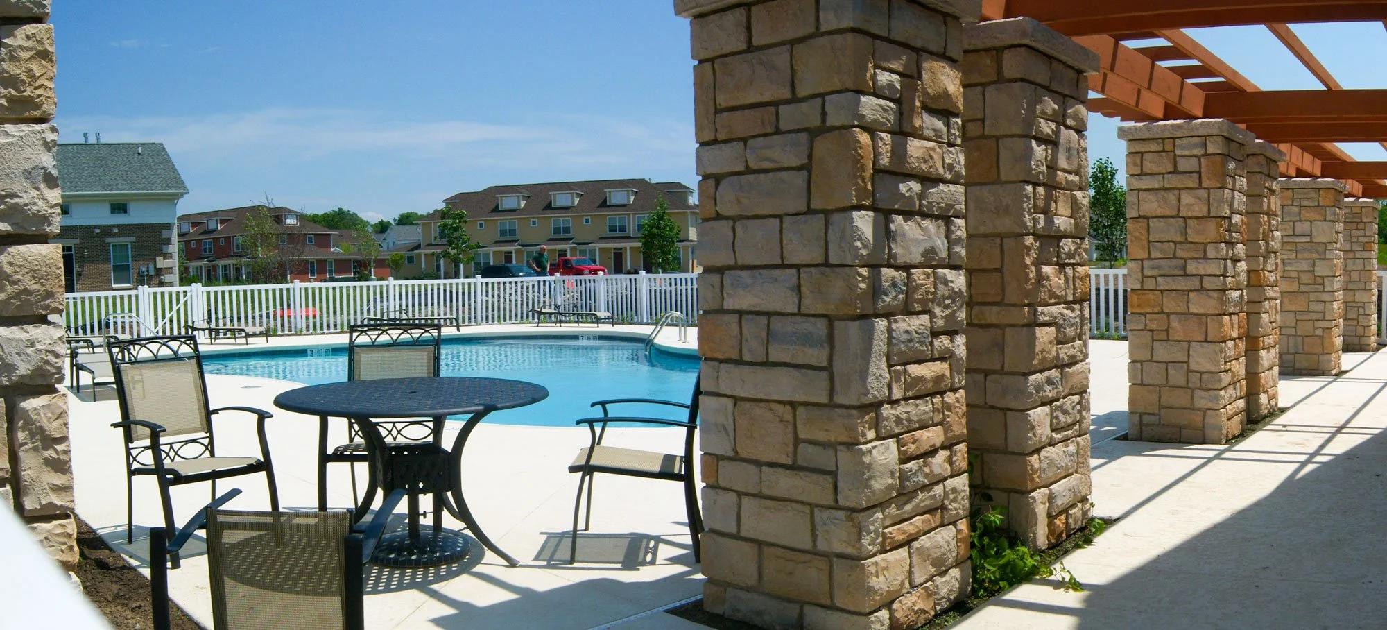 An outdoor swimming pool area with surrounding chairs and tables, stone columns supporting a pergola, and a residential neighborhood in the background with multi-story houses and a white fence.