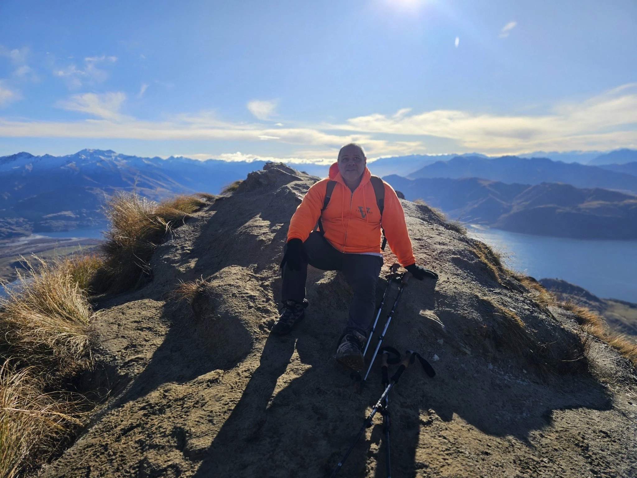 Vanguard Engineering President Joe Ardieta sitting on a rocky mountain summit with mountain range and blue sky in the background.
