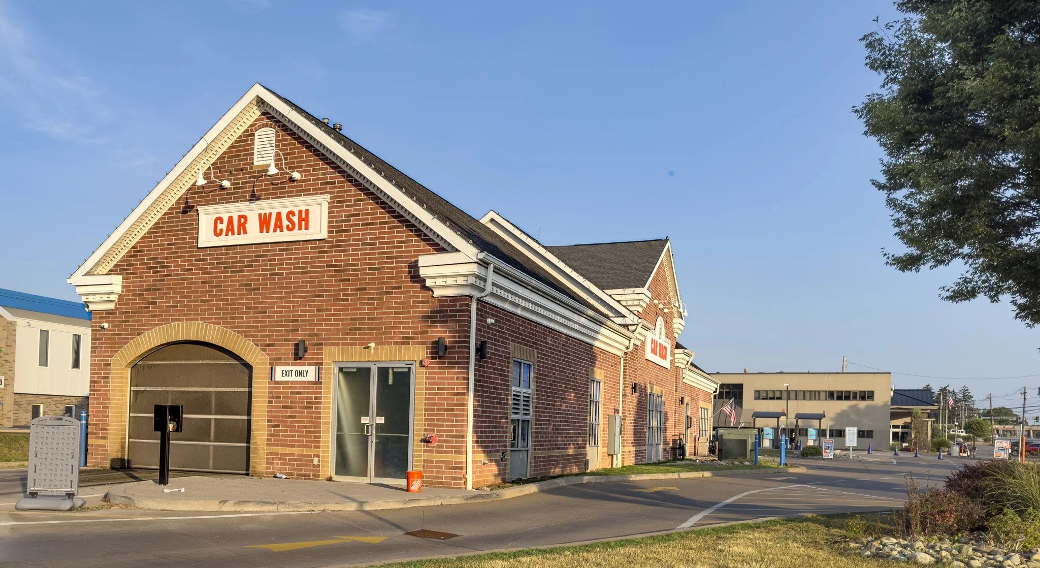 A brick building with a sign that reads 'Car Wash' on the front. The building has an arched entrance for vehicles and a glass door. There is a small sign indicating 'Exit Only' and some parking spaces in front. The sky is clear and blue, with a large tree to the right and other buildings in the background.