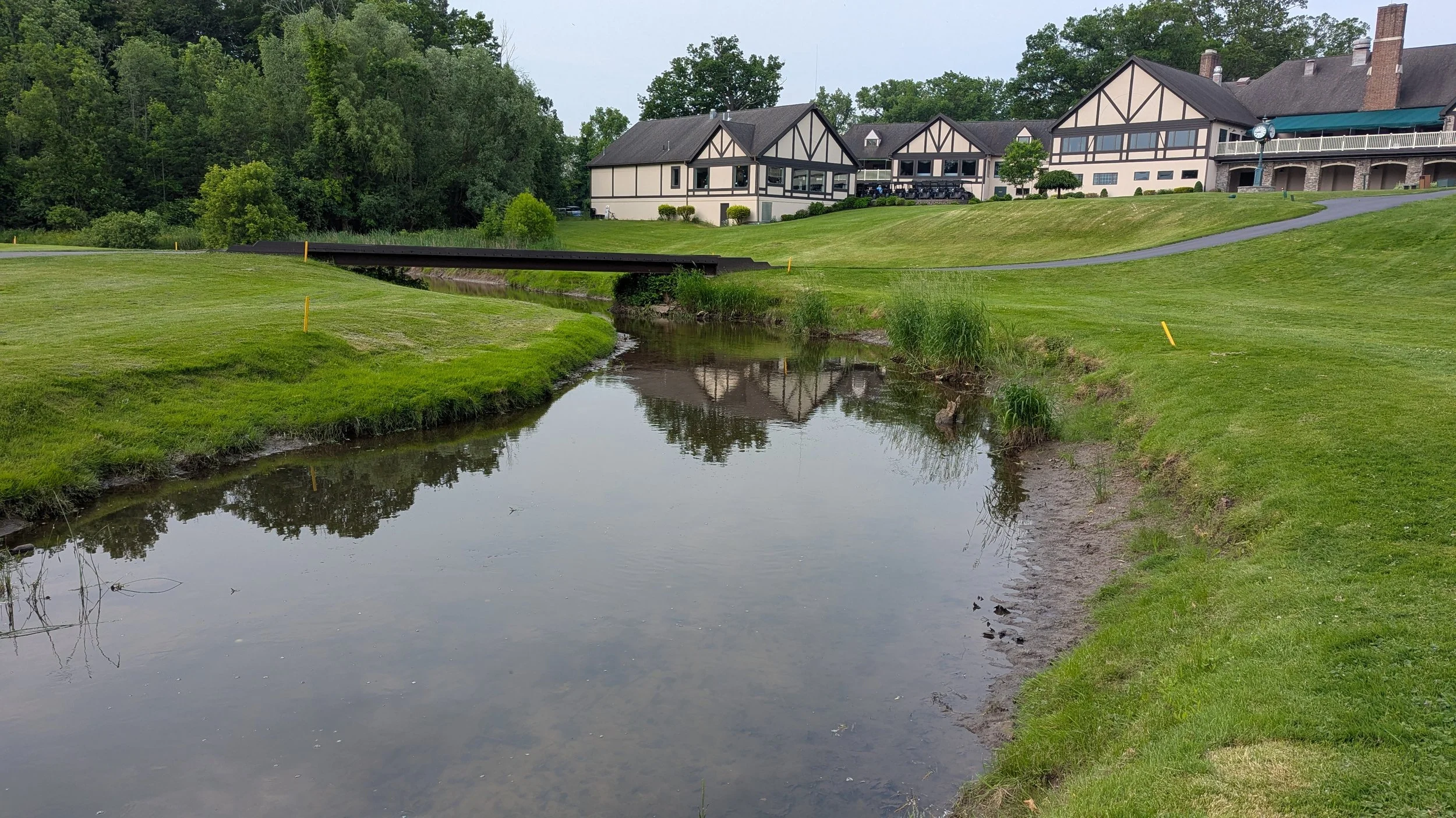 A golf course with a small creek running through it, surrounded by well-maintained grass, and a large building with multiple gabled roofs and a terrace overlooking the course in the background.
